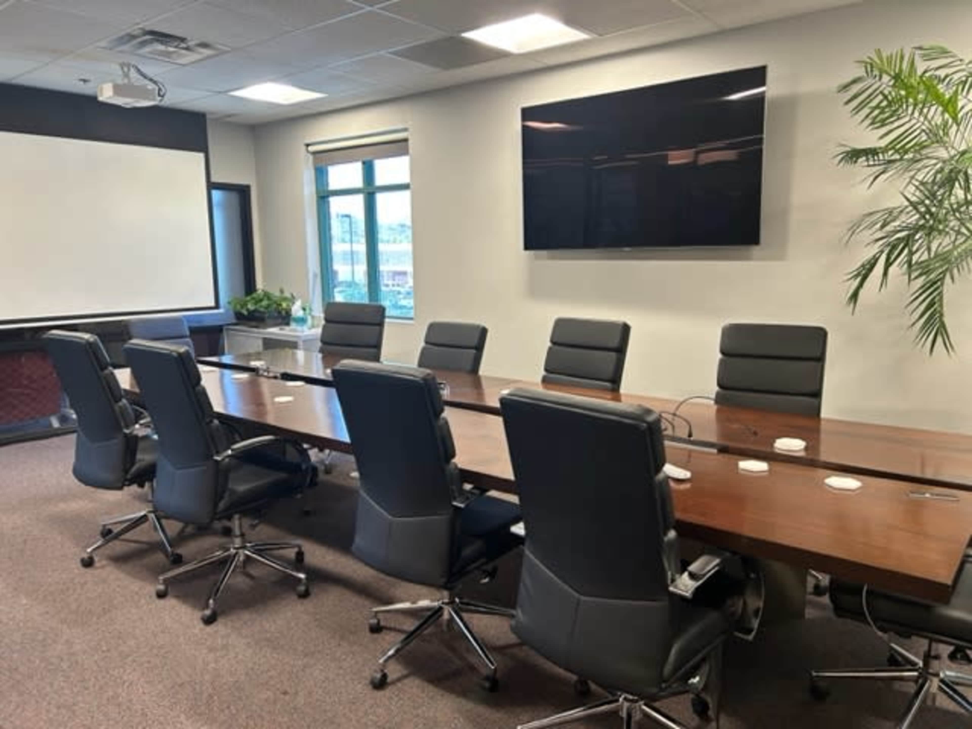 A modern conference room with a long wooden table surrounded by black ergonomic chairs, a wall-mounted screen, and large windows letting in natural light.