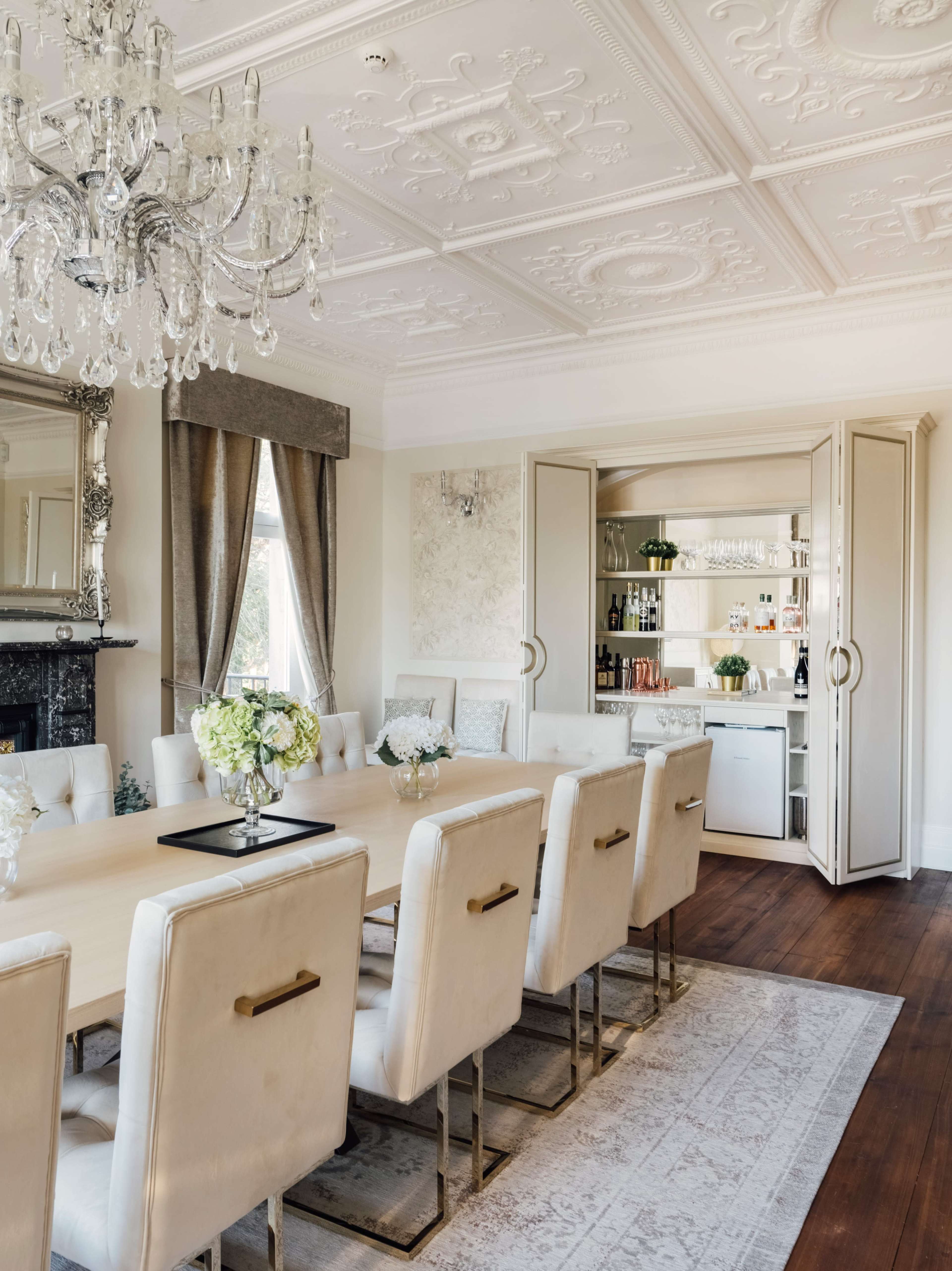 The dining room features a long table surrounded by upholstered chairs, with a chandelier hanging from a decorative ceiling and an adjacent cabinet showcasing various drinks and glassware.
