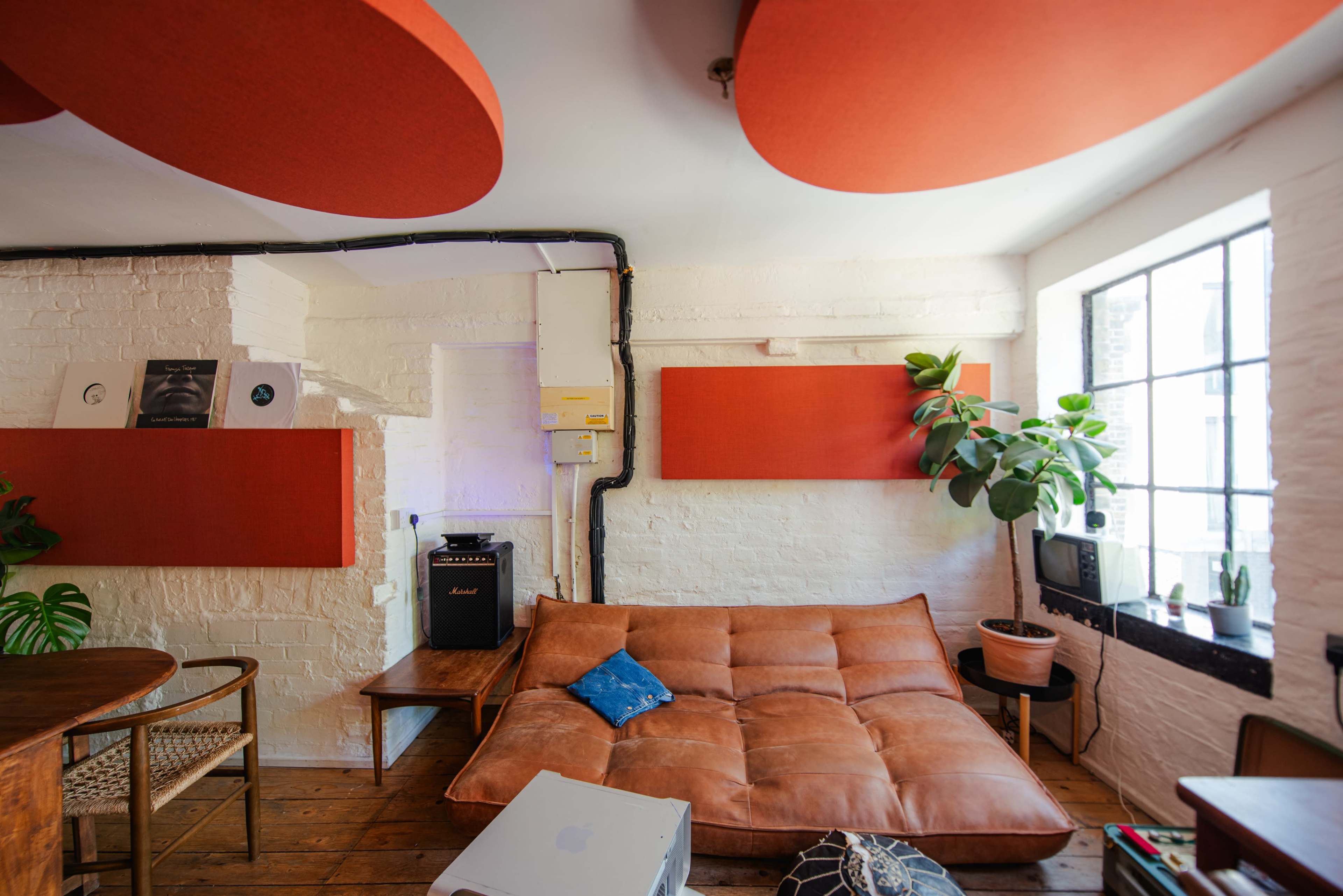 The image shows a cozy room with a large brown couch, wooden flooring, a small plant, and sound equipment on display next to a window.
