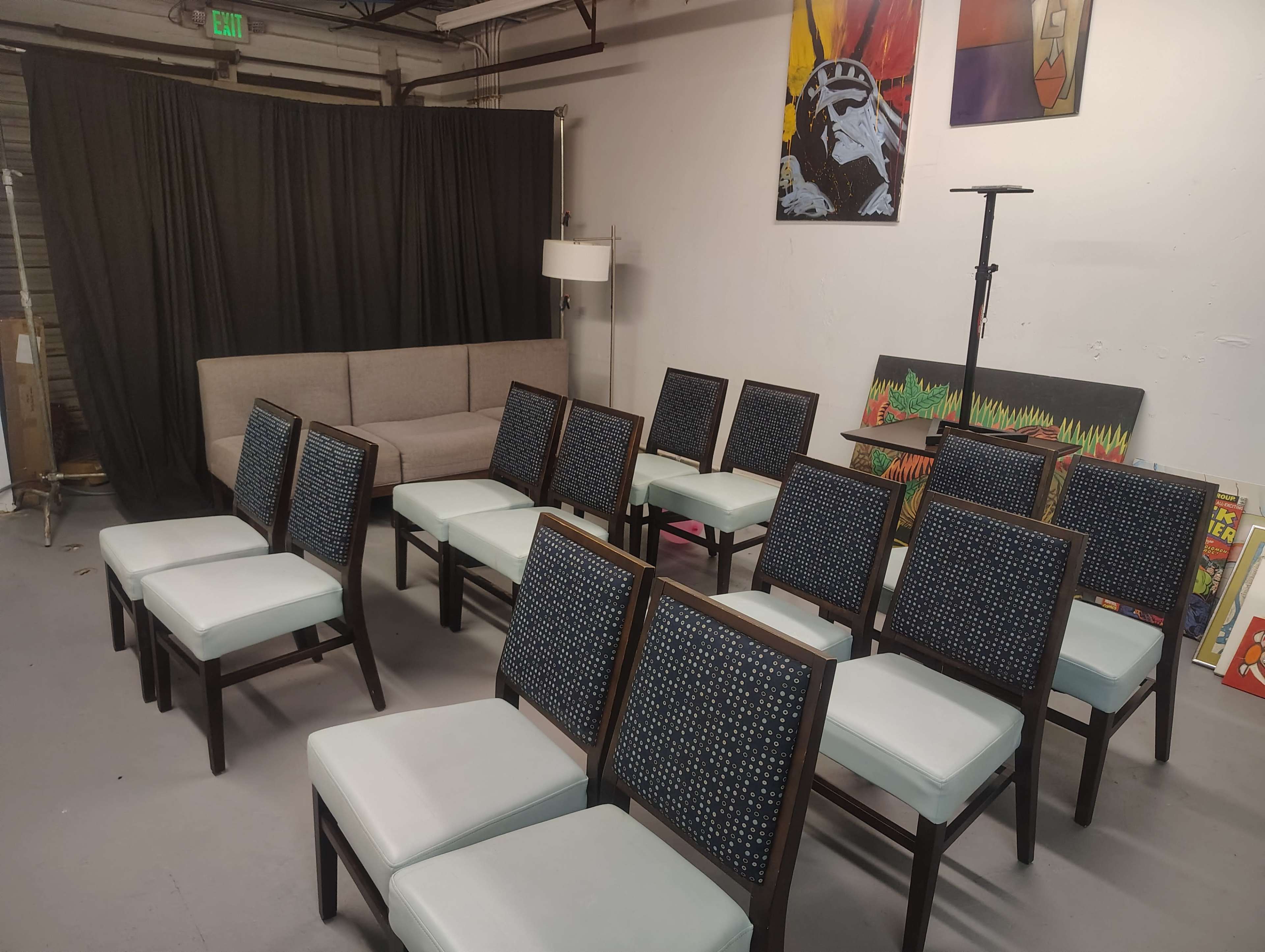 The image shows a room arranged with a gray sofa, a set of twelve wooden chairs with light blue cushions, and various artworks displayed on the walls.