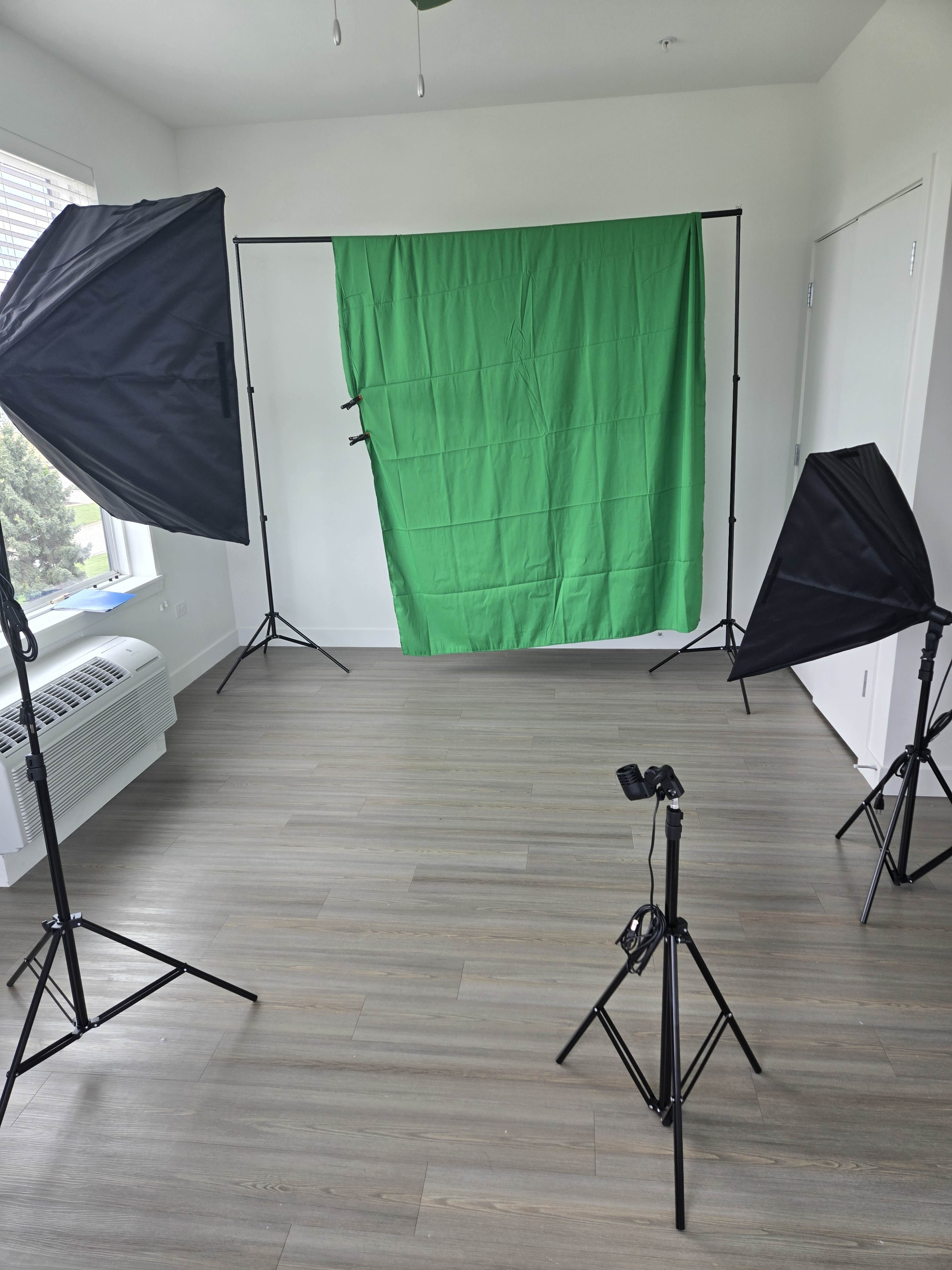 The image shows a photography set up in a room with a green backdrop, two softbox lights, and a tripod with a camera.