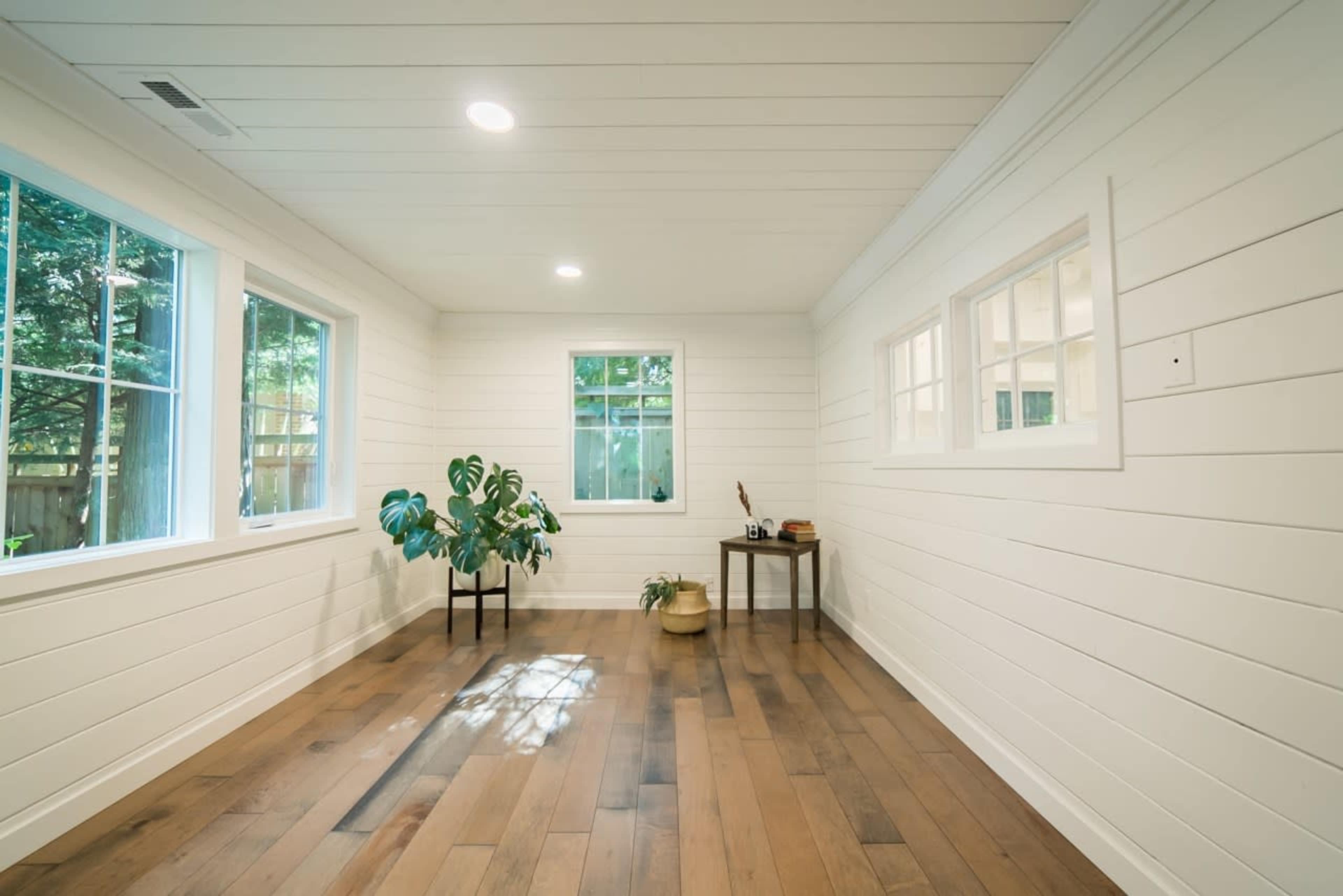 A bright, empty room with white shiplap walls and large windows, featuring hardwood floors and a small plant in the corner.