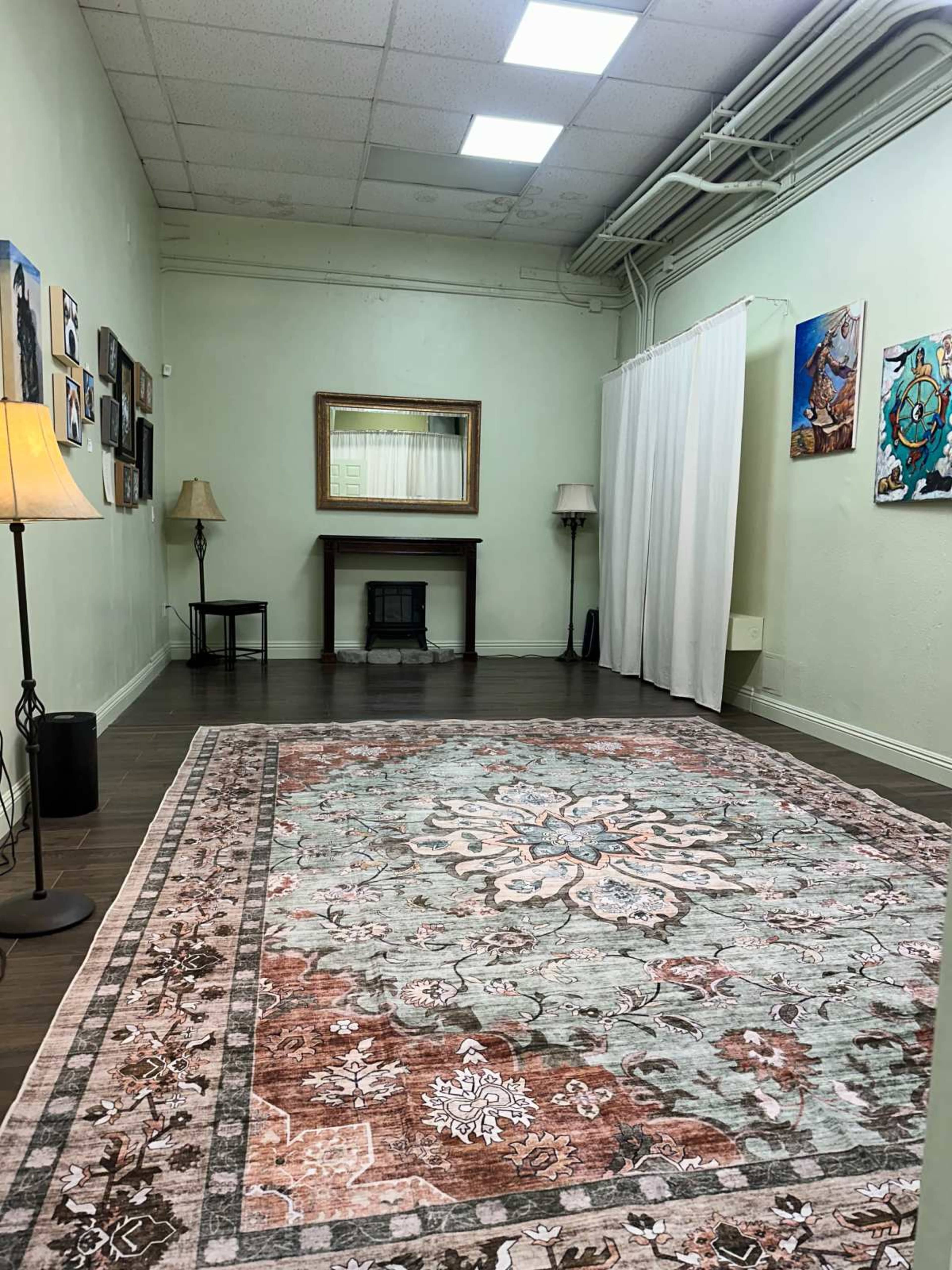 The image shows a spacious room with a large decorative rug, two lamps, a small table with a mirror above it, and wall art displayed on the light green walls.