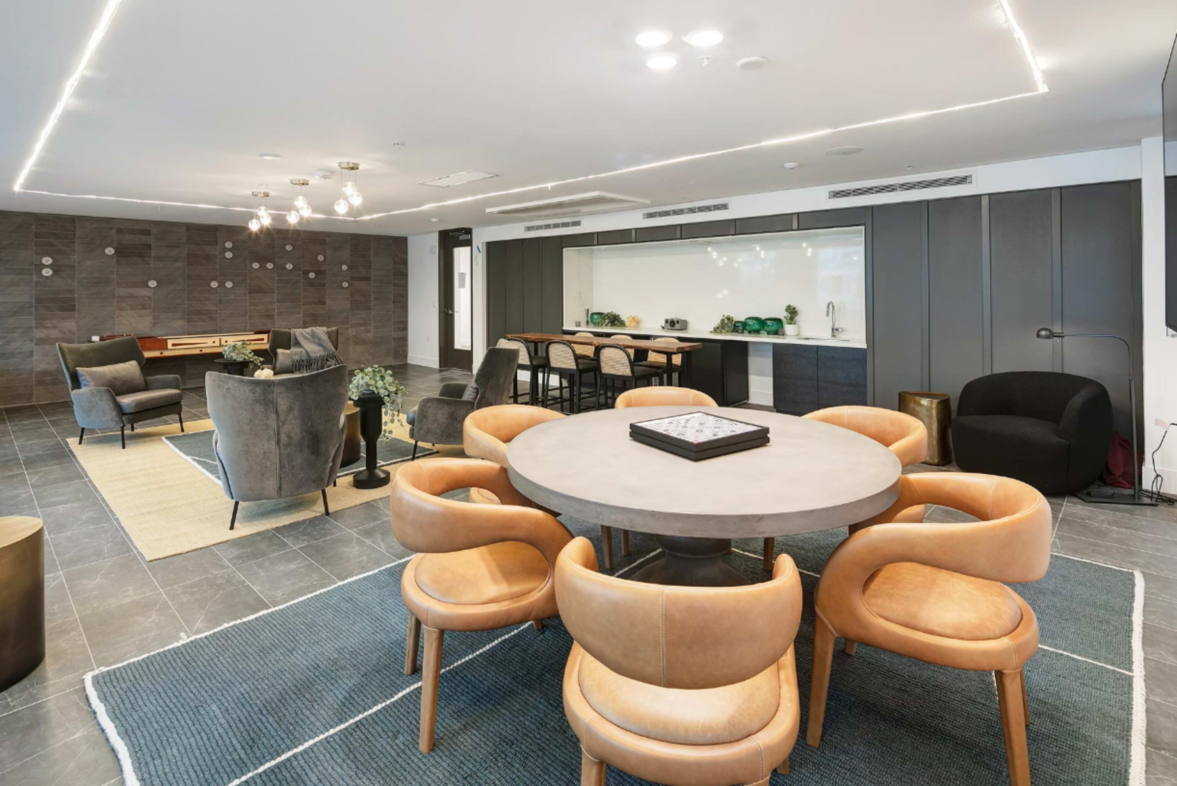 A modern dining area features a round table surrounded by eight tan chairs, with a sleek kitchen and lounge space in the background.