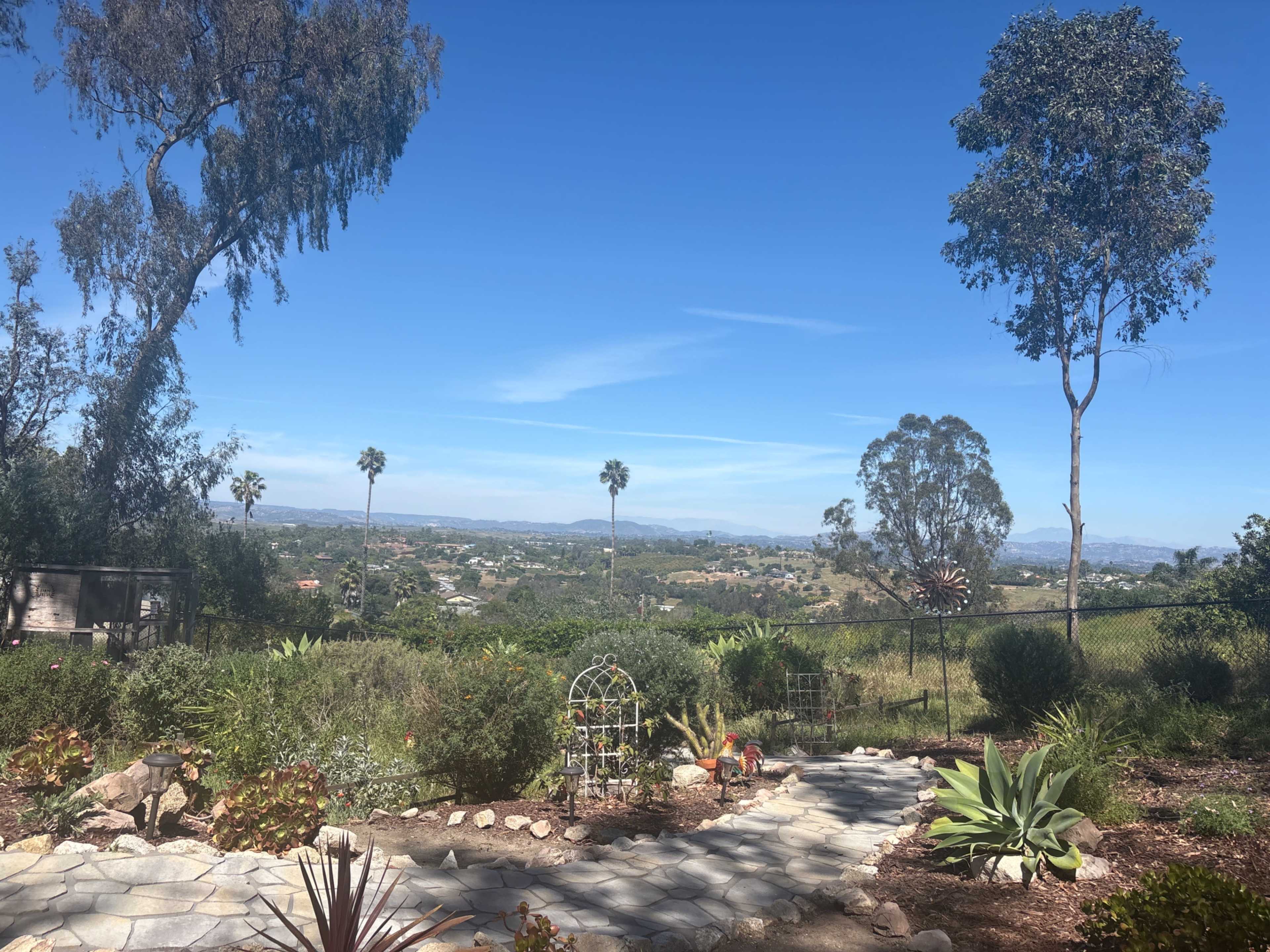 The image shows a landscaped garden with a stone pathway leading to a view of hills and distant palm trees under a clear blue sky.