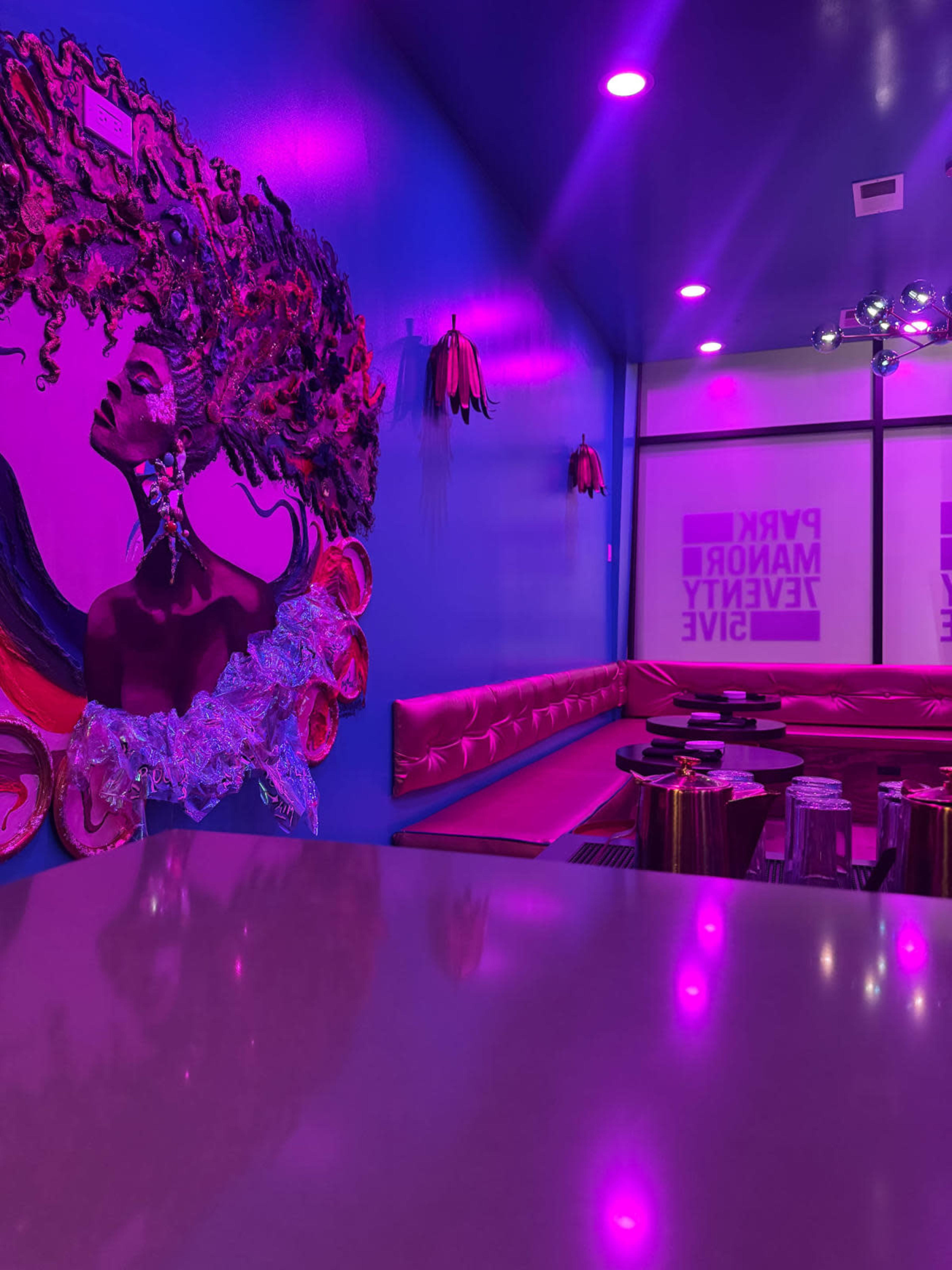 The image shows a vibrant room with purple lighting, featuring a large artistic mural of a woman's profile on a blue wall and a stylish seating area with a glossy countertop.