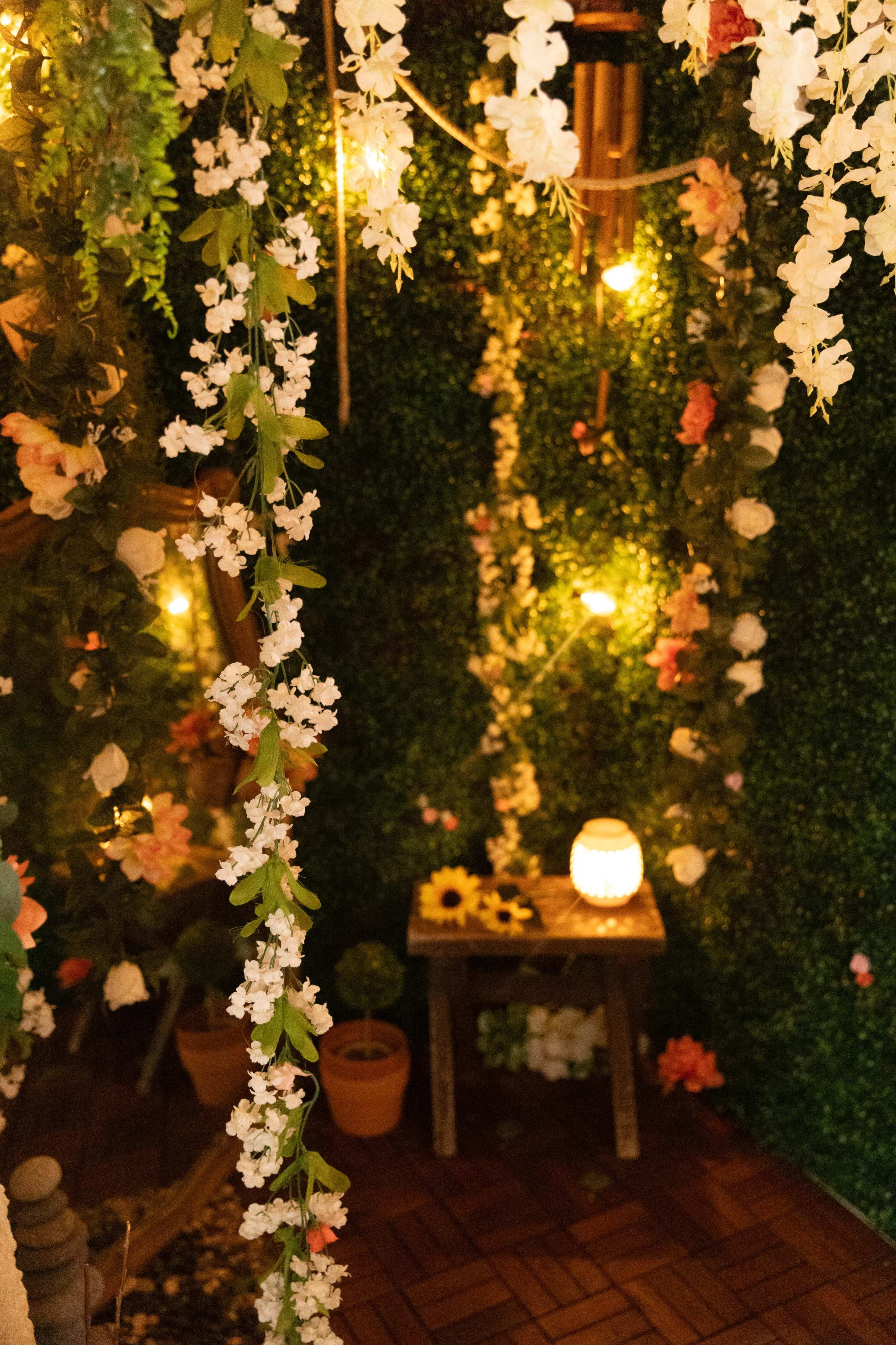 A decorative indoor space filled with artificial flowers, string lights, and a small table with a lantern and potted plants.