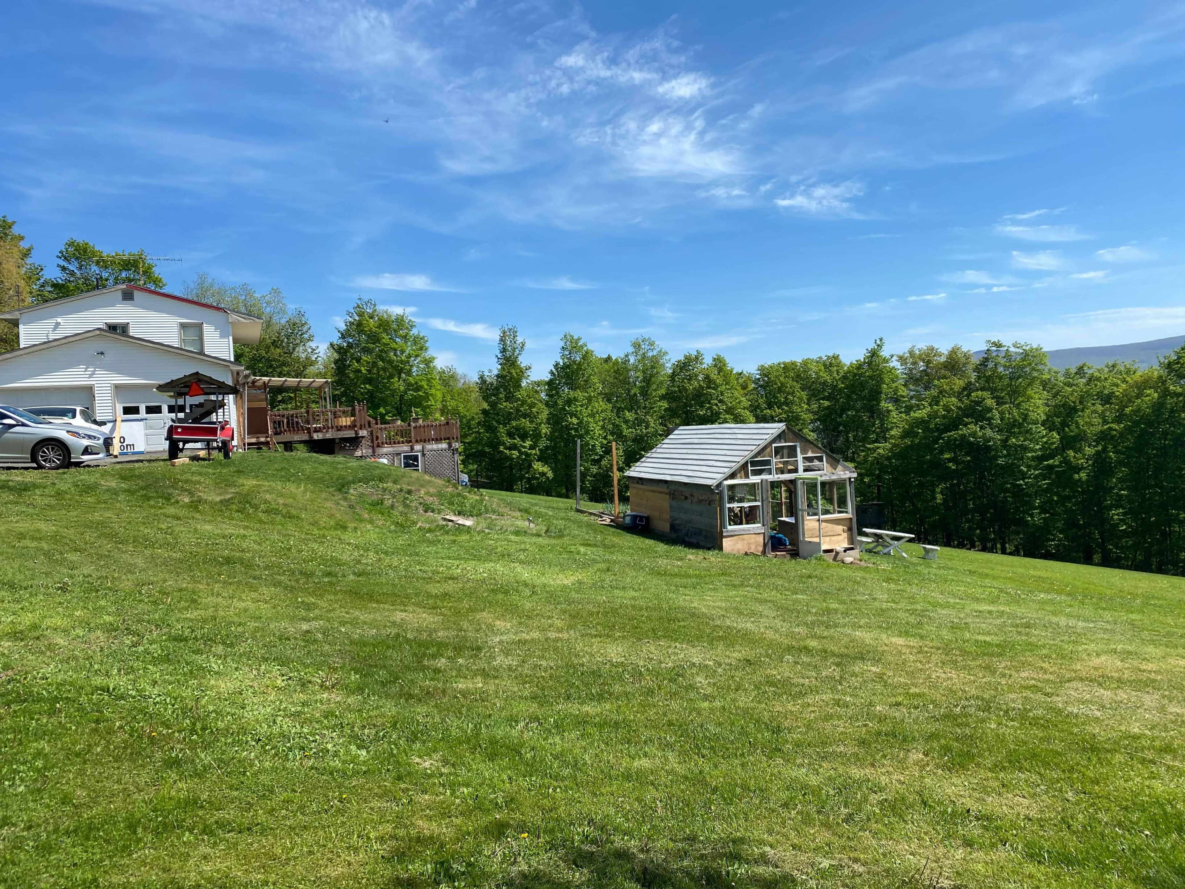 A house with a deck and a greenhouse sits on a grassy hillside surrounded by trees and blue sky.