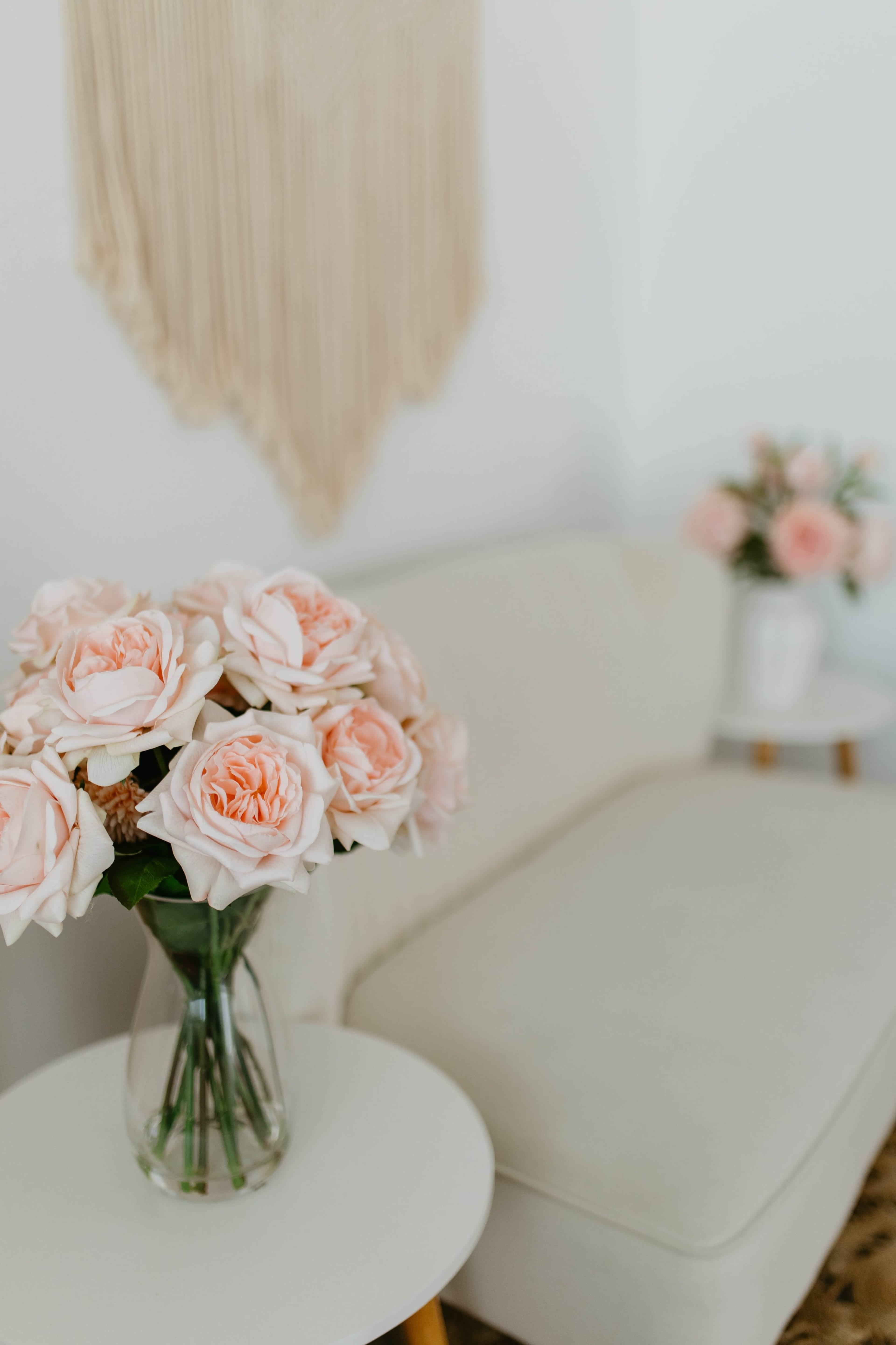 A vase of pink roses sits on a small white table next to a beige sofa in a minimally decorated room.