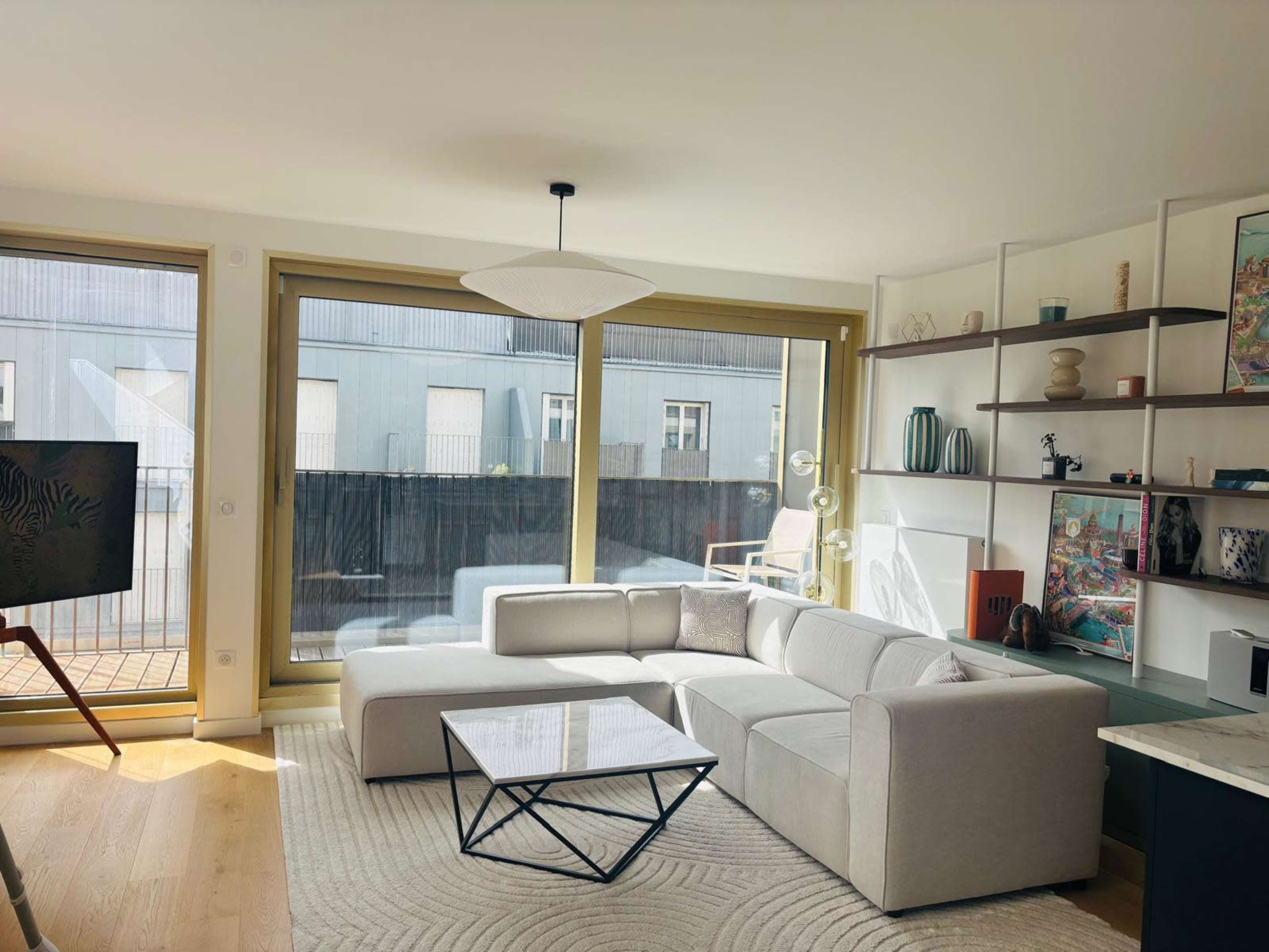 The image shows a modern living room with large windows, a sectional sofa, a glass coffee table, and shelves displaying various decorative items.