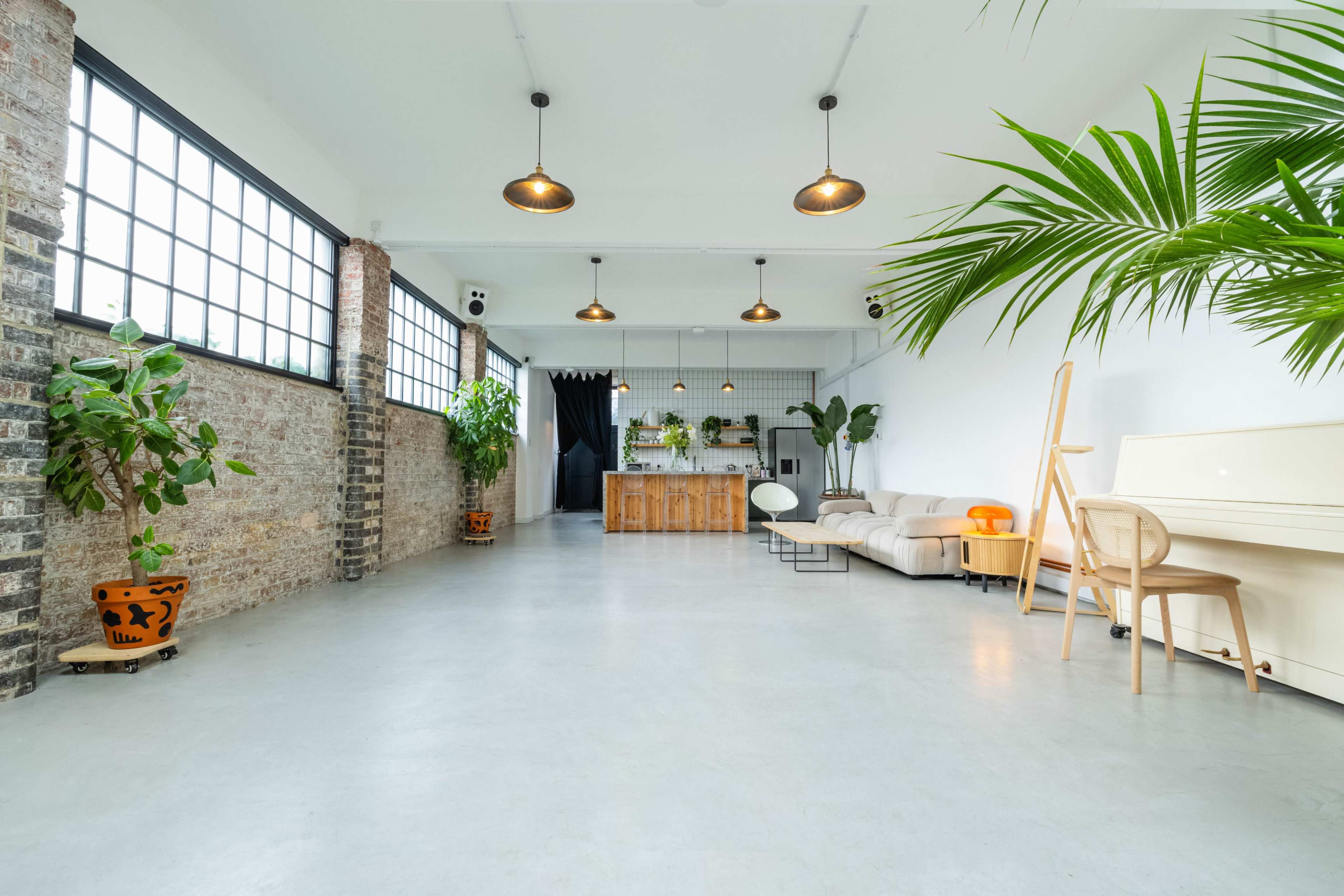 A spacious interior with large windows, wooden accents, plants, and a light-colored sofa, leading to a kitchen area.