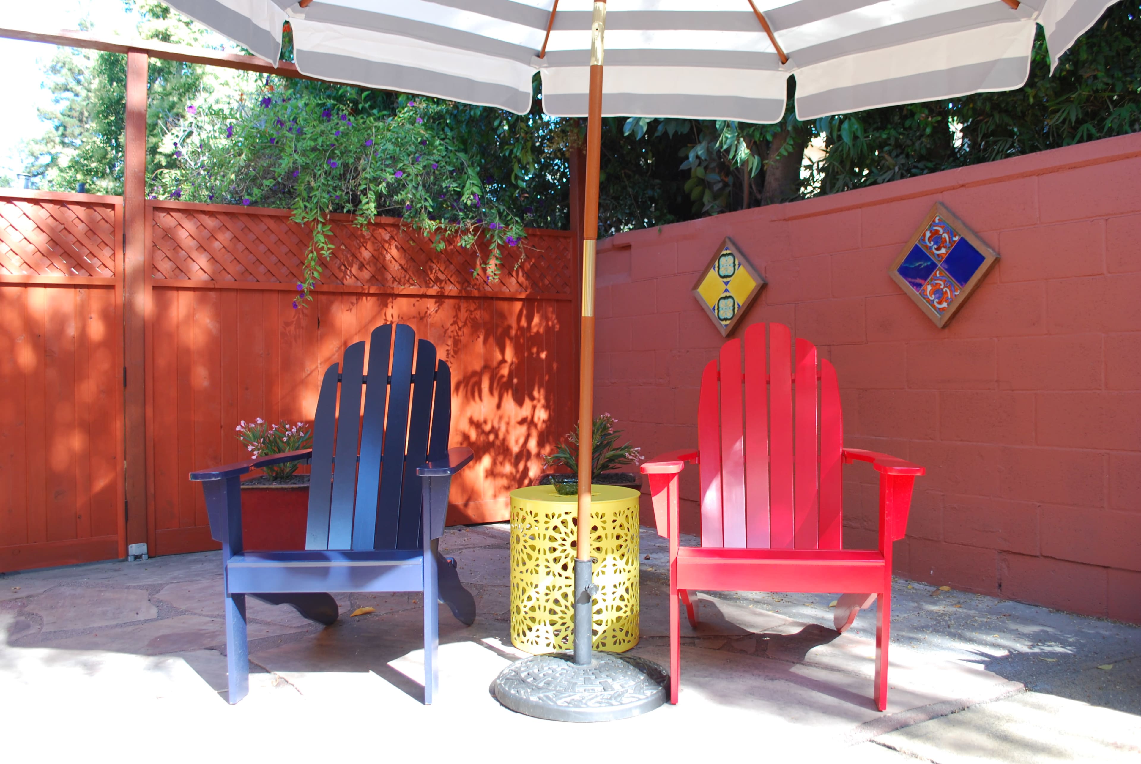 Two adirondack chairs, one navy blue and one bright red, under a striped umbrella on a patio surrounded by a terracotta-colored wall and decorative plants.