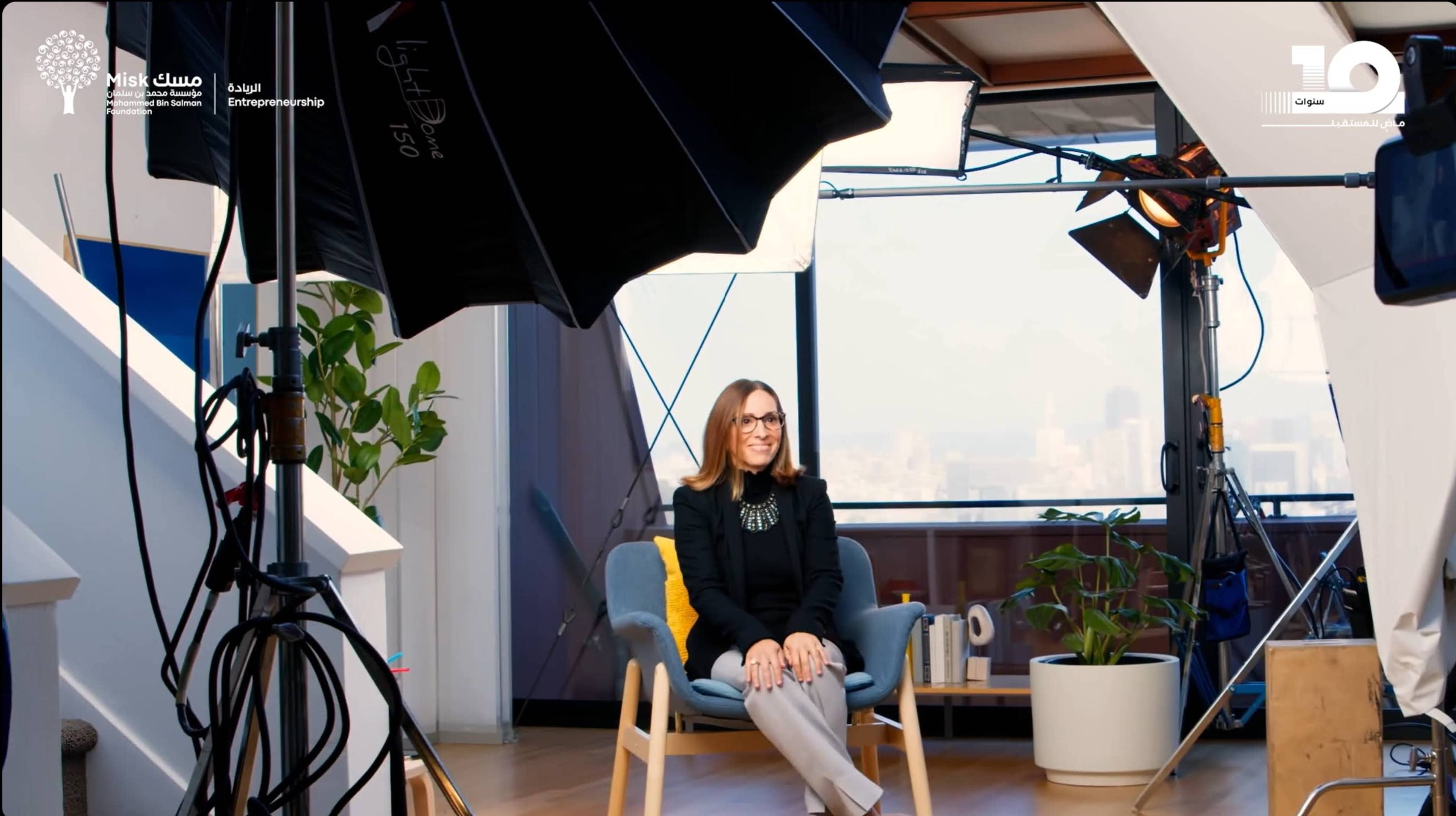 A woman sits on a chair in a well-lit studio with large softbox lights and a window showing a city view in the background.