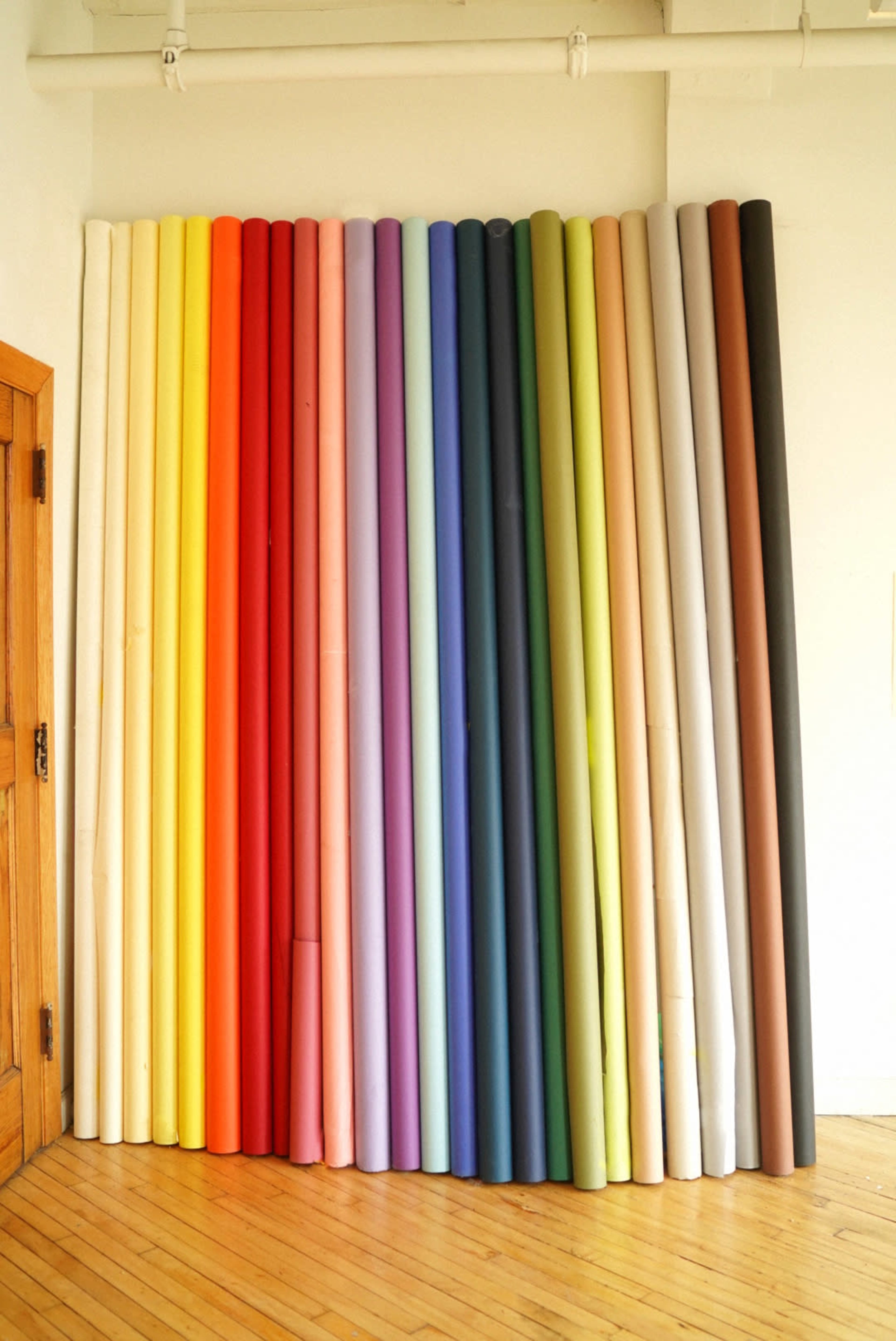The image shows a collection of large rolls of colored paper arranged vertically against a wall.