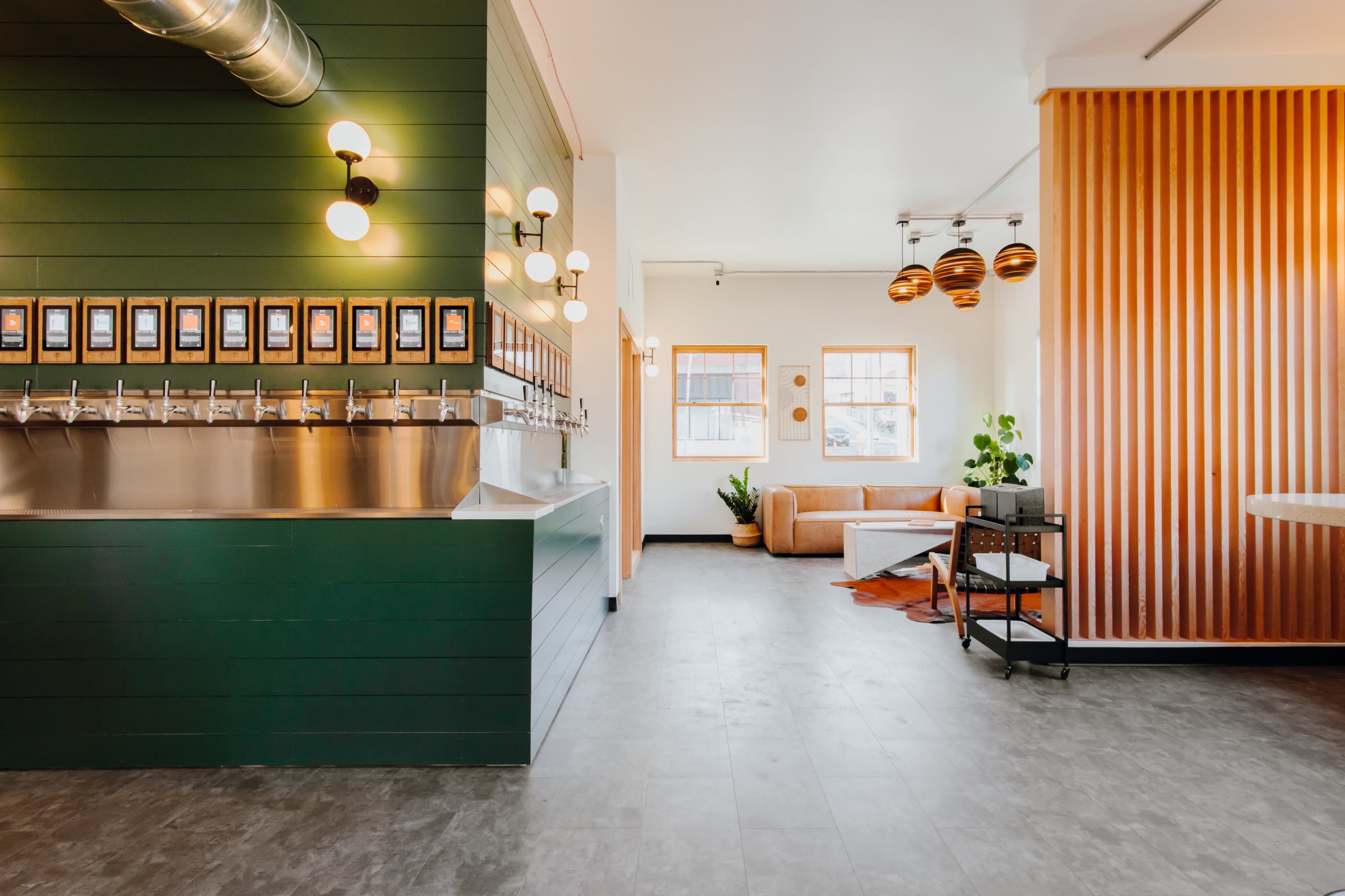 A modern interior with a green wall featuring multiple drink taps on one side and a cozy seating area with wooden accents and plants on the other.