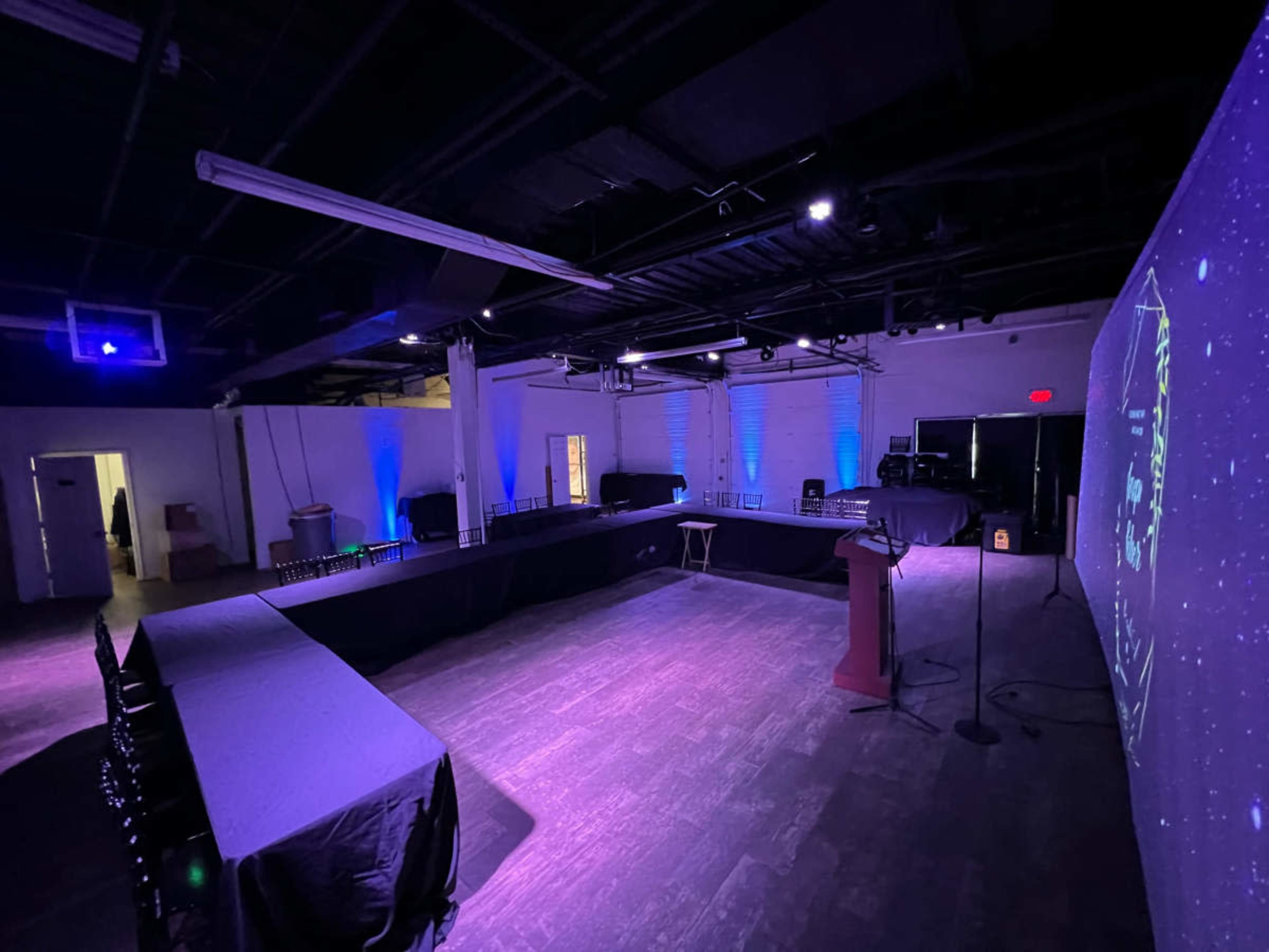The image shows a dimly lit event space with multiple tables arranged in a U-shape, accompanied by a podium and a screen displaying a presentation.