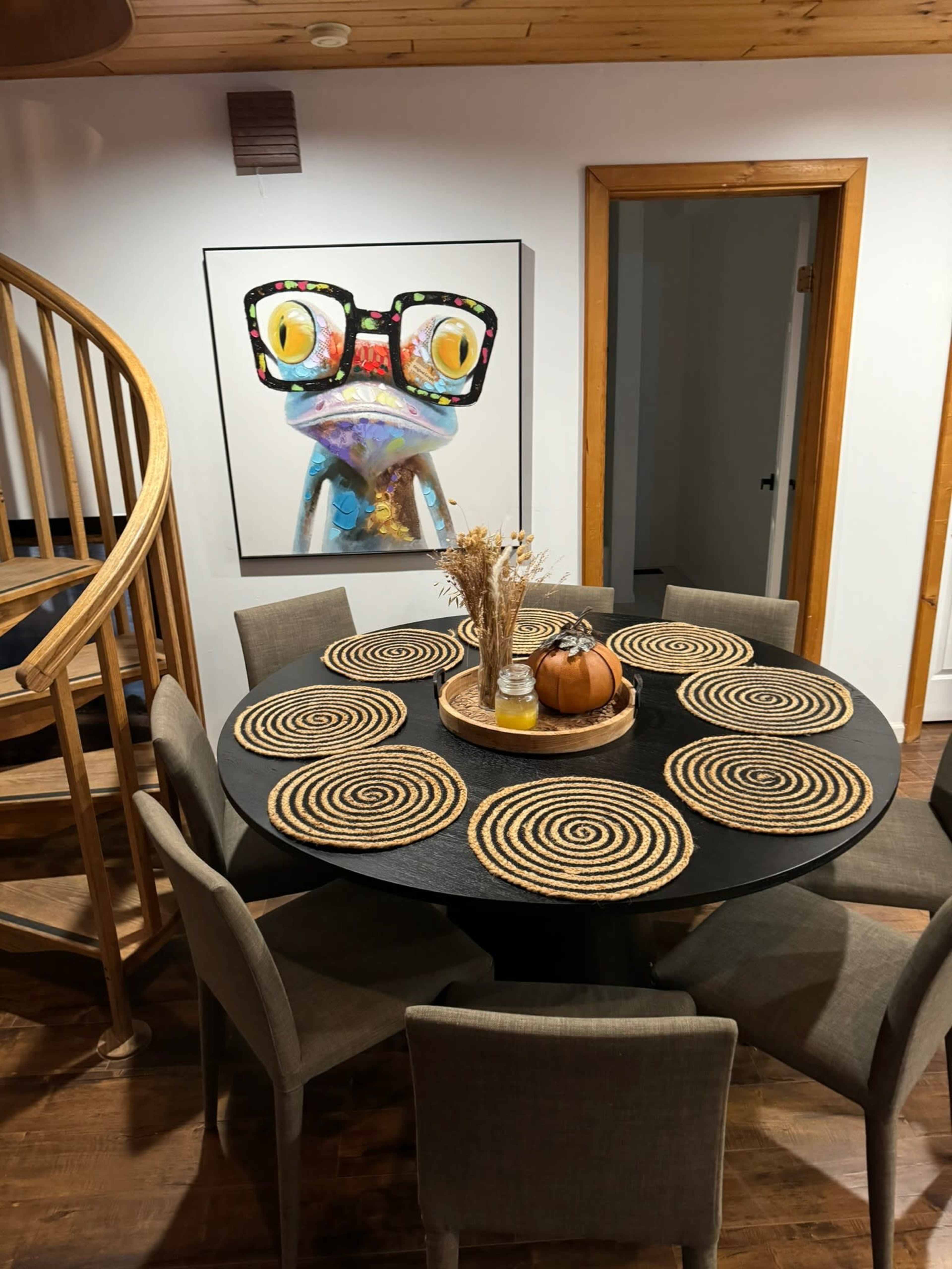 A round dining table with woven placemats is set against a backdrop featuring a large colorful frog artwork and a spiral staircase.