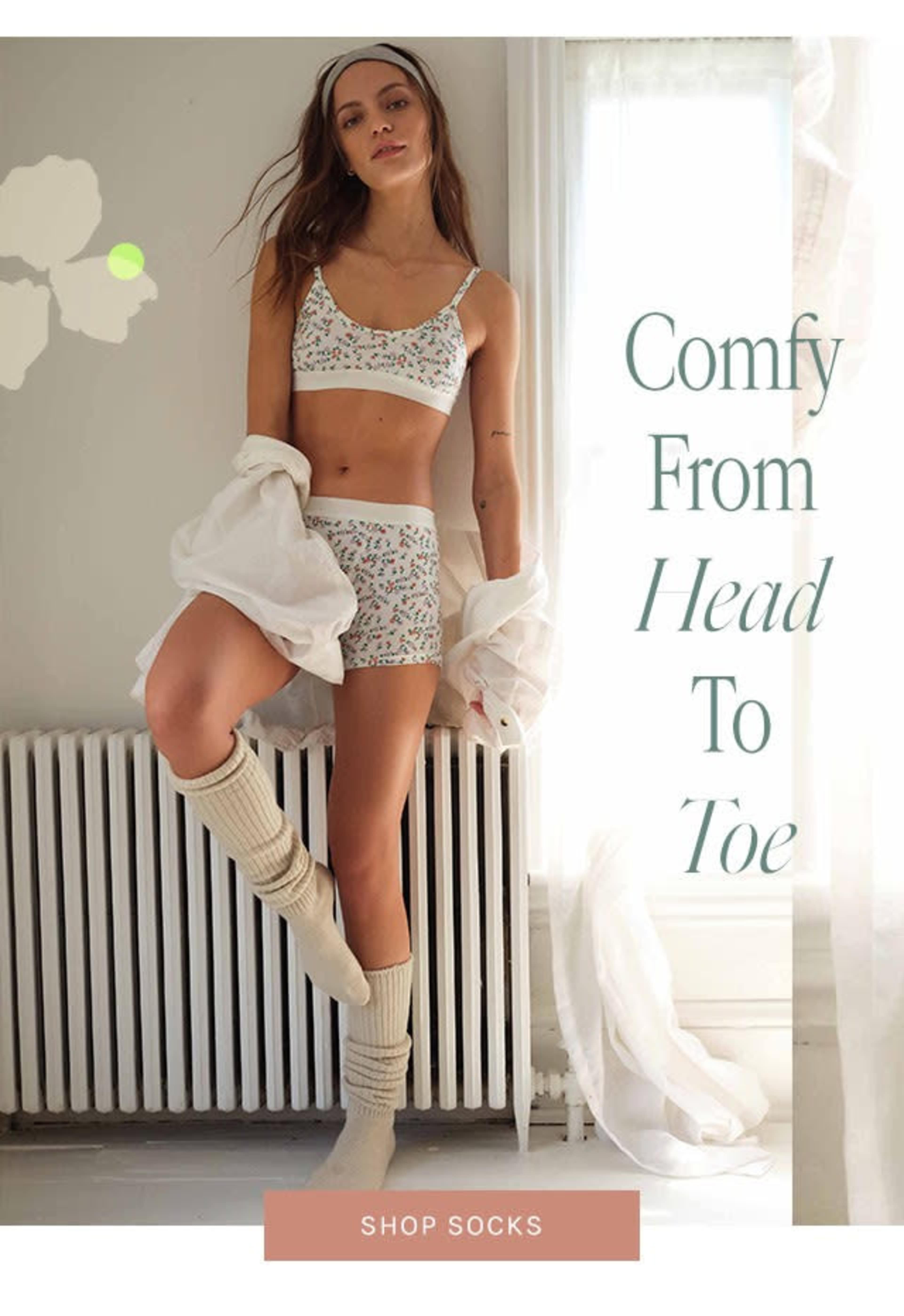 A model poses in a floral lingerie set and knee-high socks next to a radiator in a bright room.
