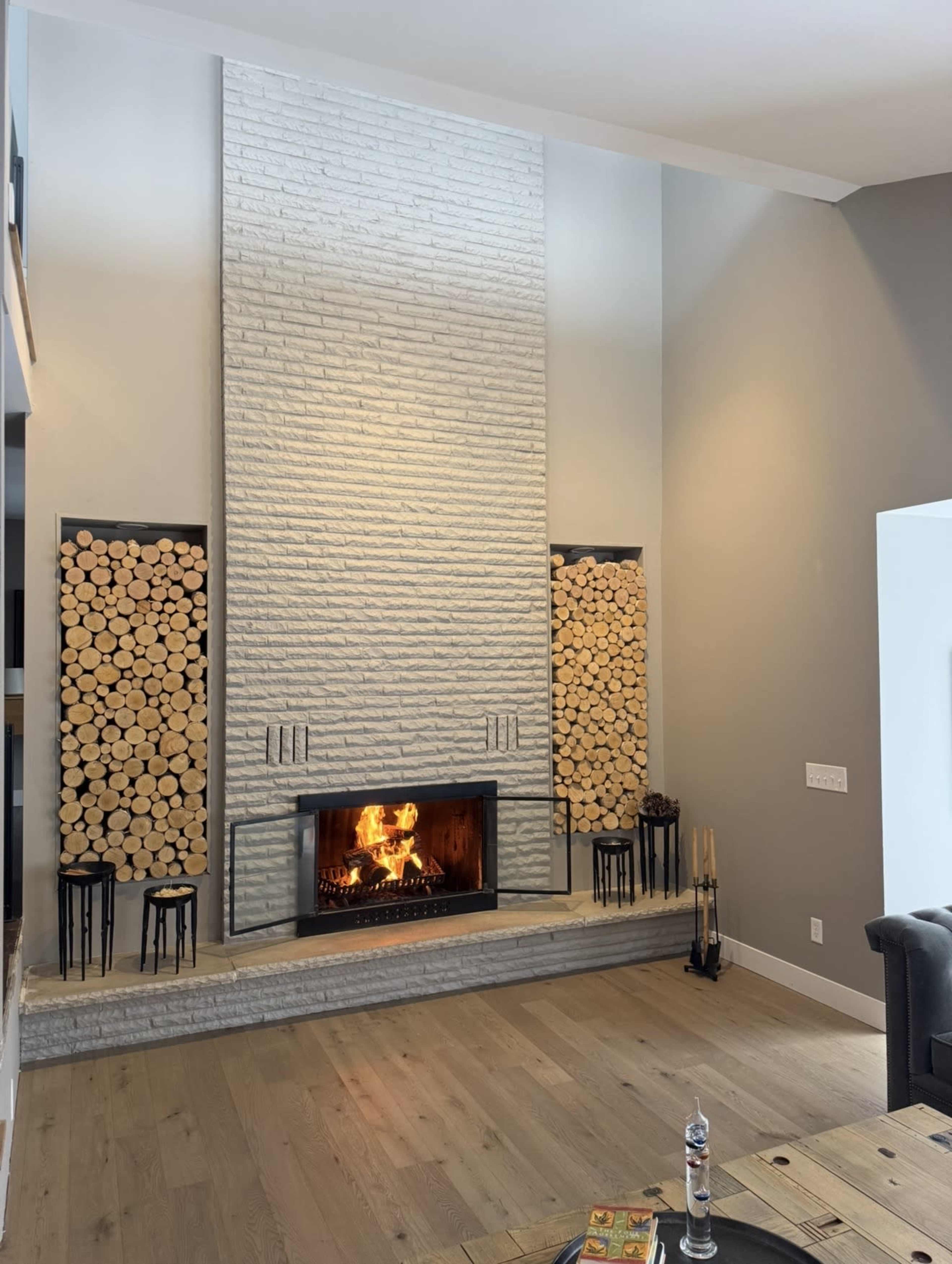 The image shows a modern living room featuring a large stone fireplace with a visible fire, flanked by two stacks of firewood and minimalistic black stools.