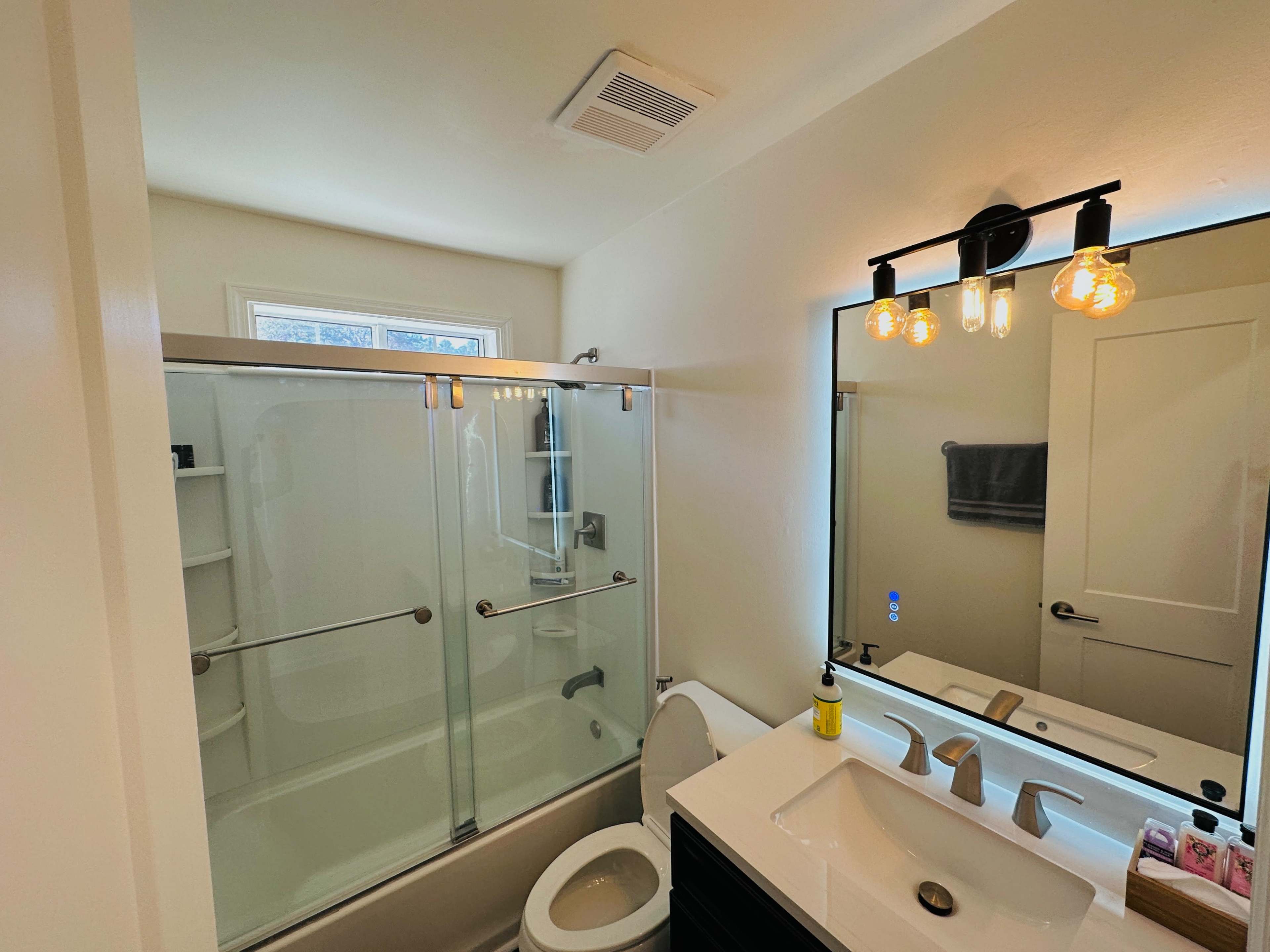 A modern bathroom with a glass shower enclosure, a white sink with two faucets, a toilet, and a large mirror with three light bulbs above it.
