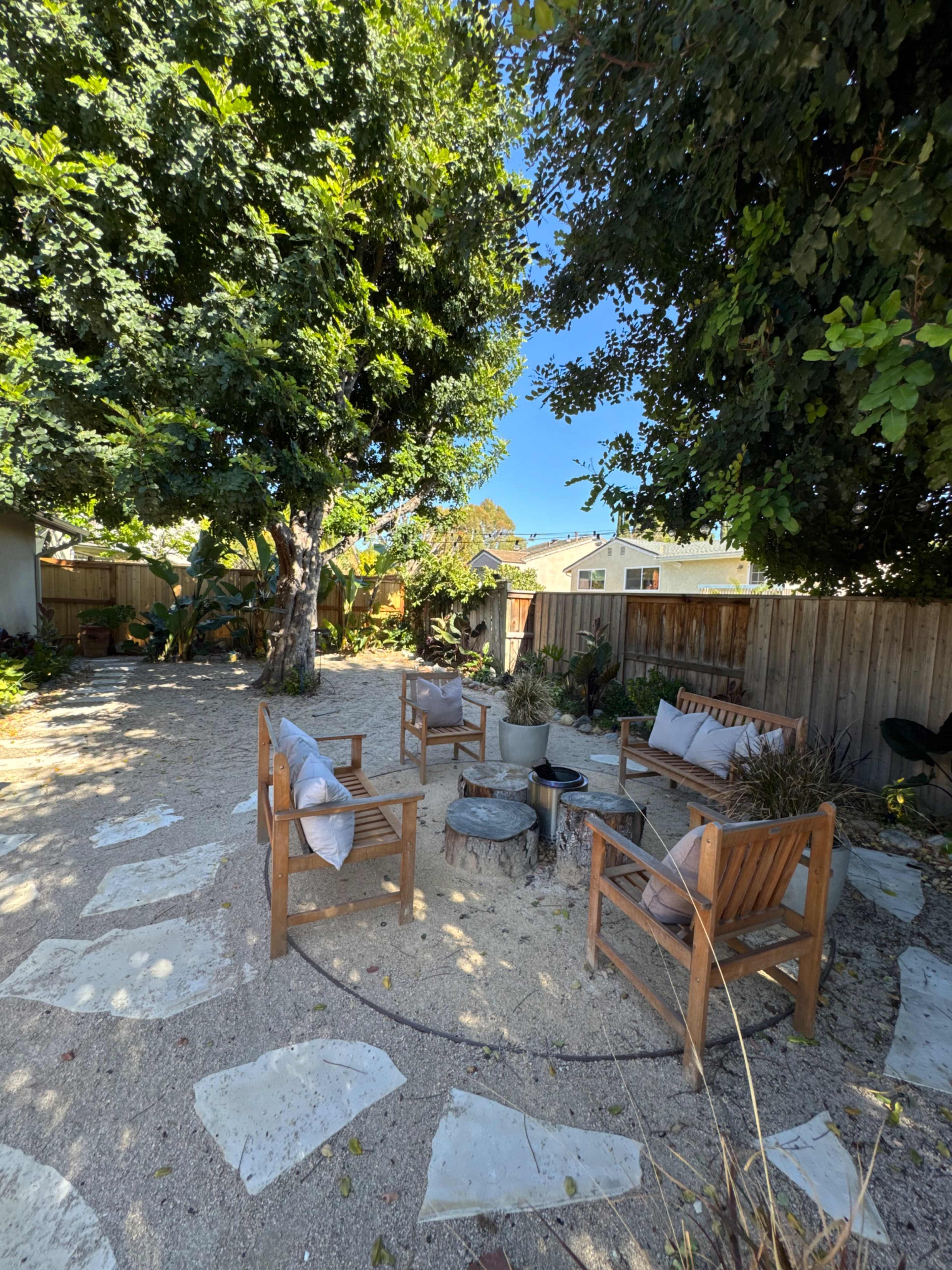 The image shows a landscaped backyard featuring a circular seating area with wooden chairs surrounding a small fire pit, set among gravel and stone pathways.