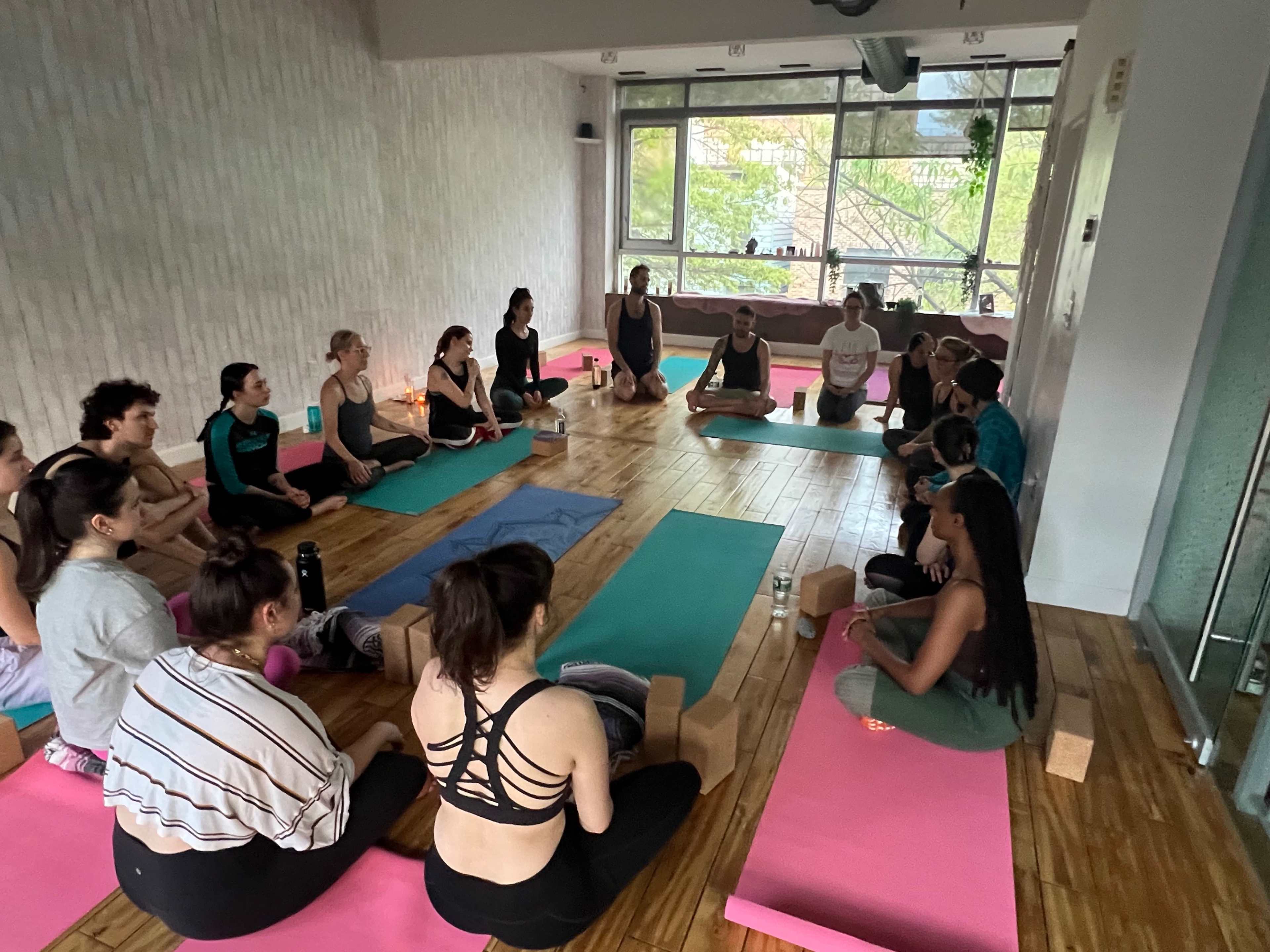 A group of people practices yoga in a studio featuring wooden floors and large windows overlooking greenery.