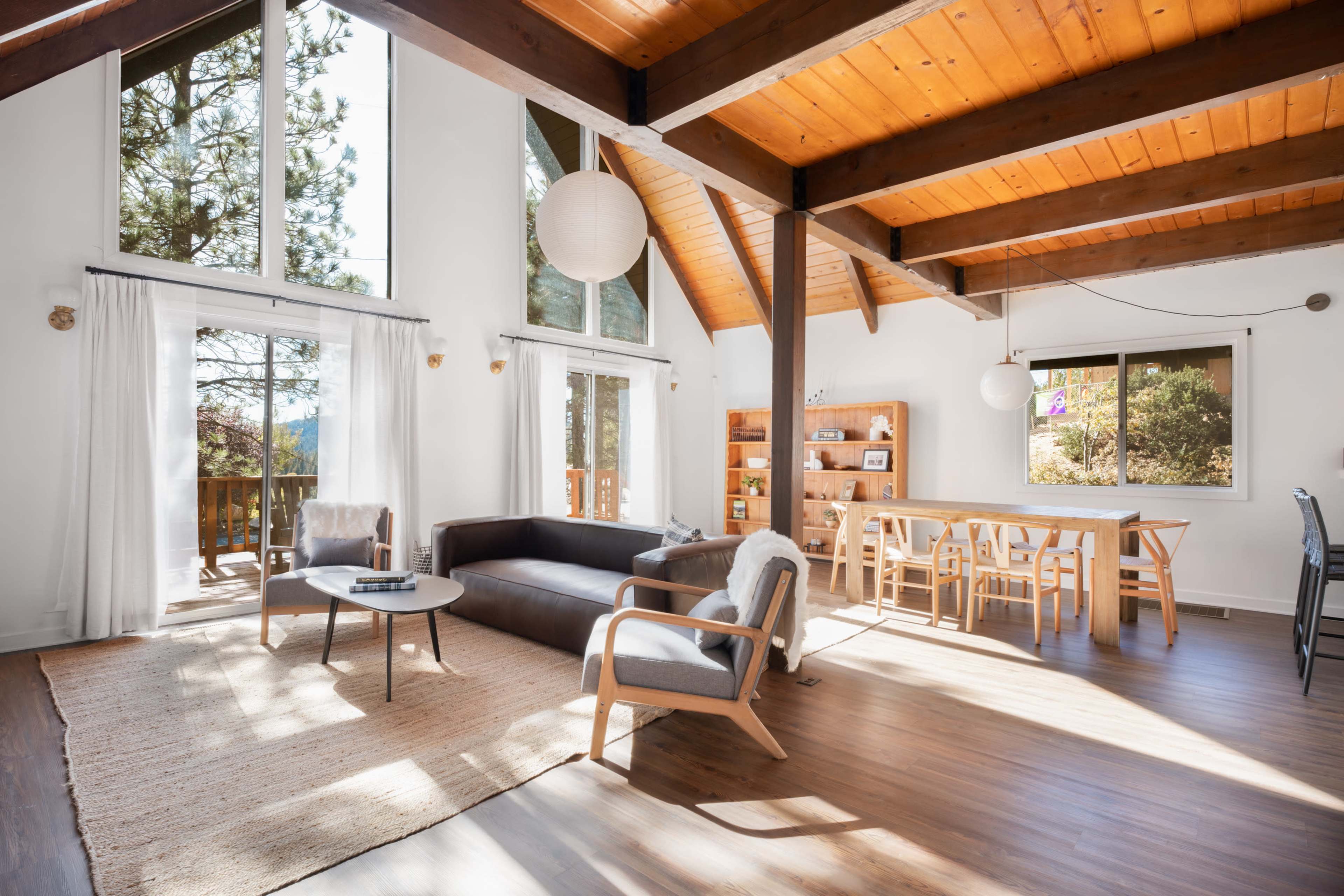 The image shows a bright, open living space with a sectional couch, dining area, and large windows showcasing a view of trees outside.