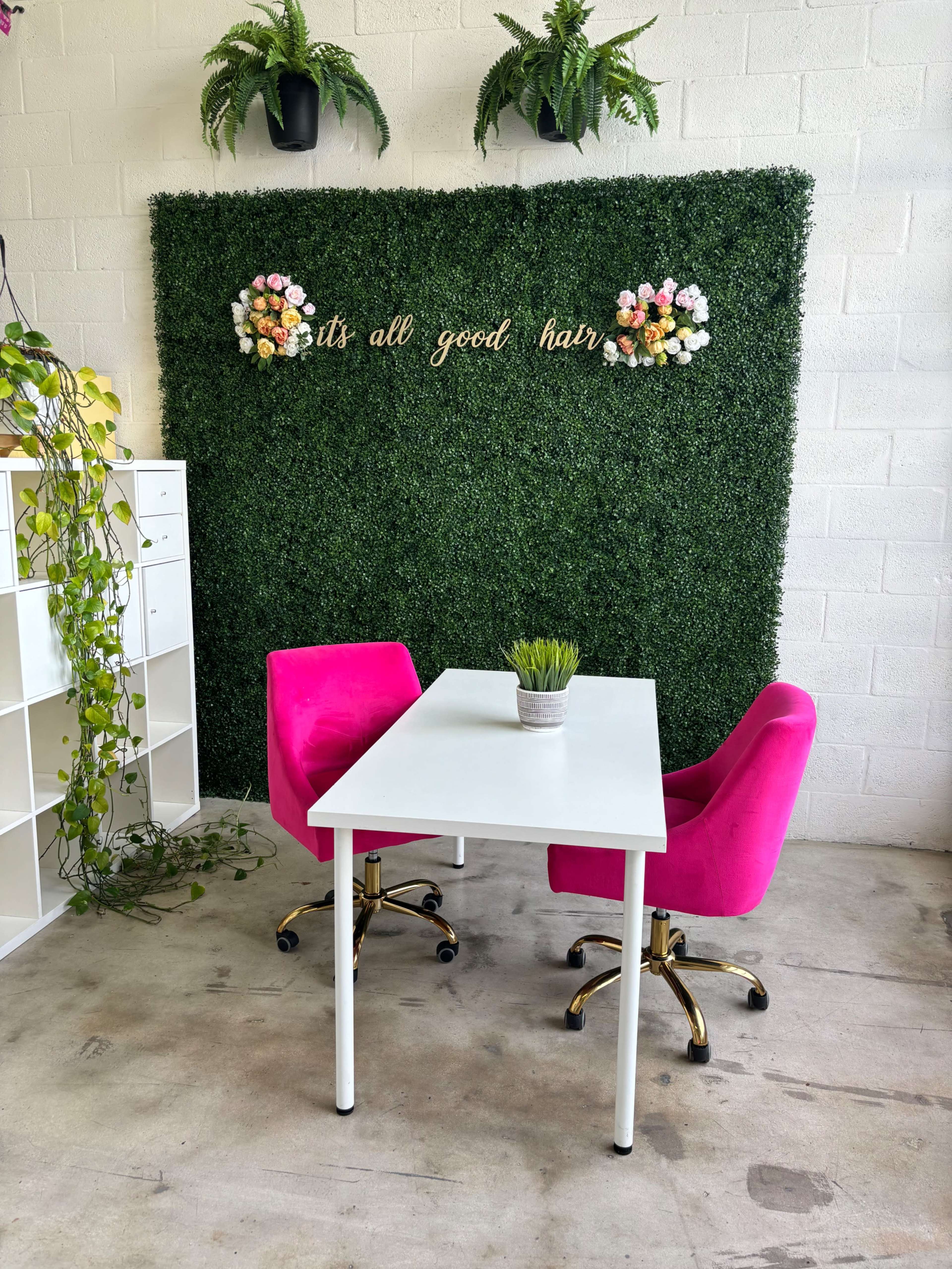 A small white table with two pink velvet chairs sits in front of a green wall adorned with artificial flowers and the text "it's all good hair."