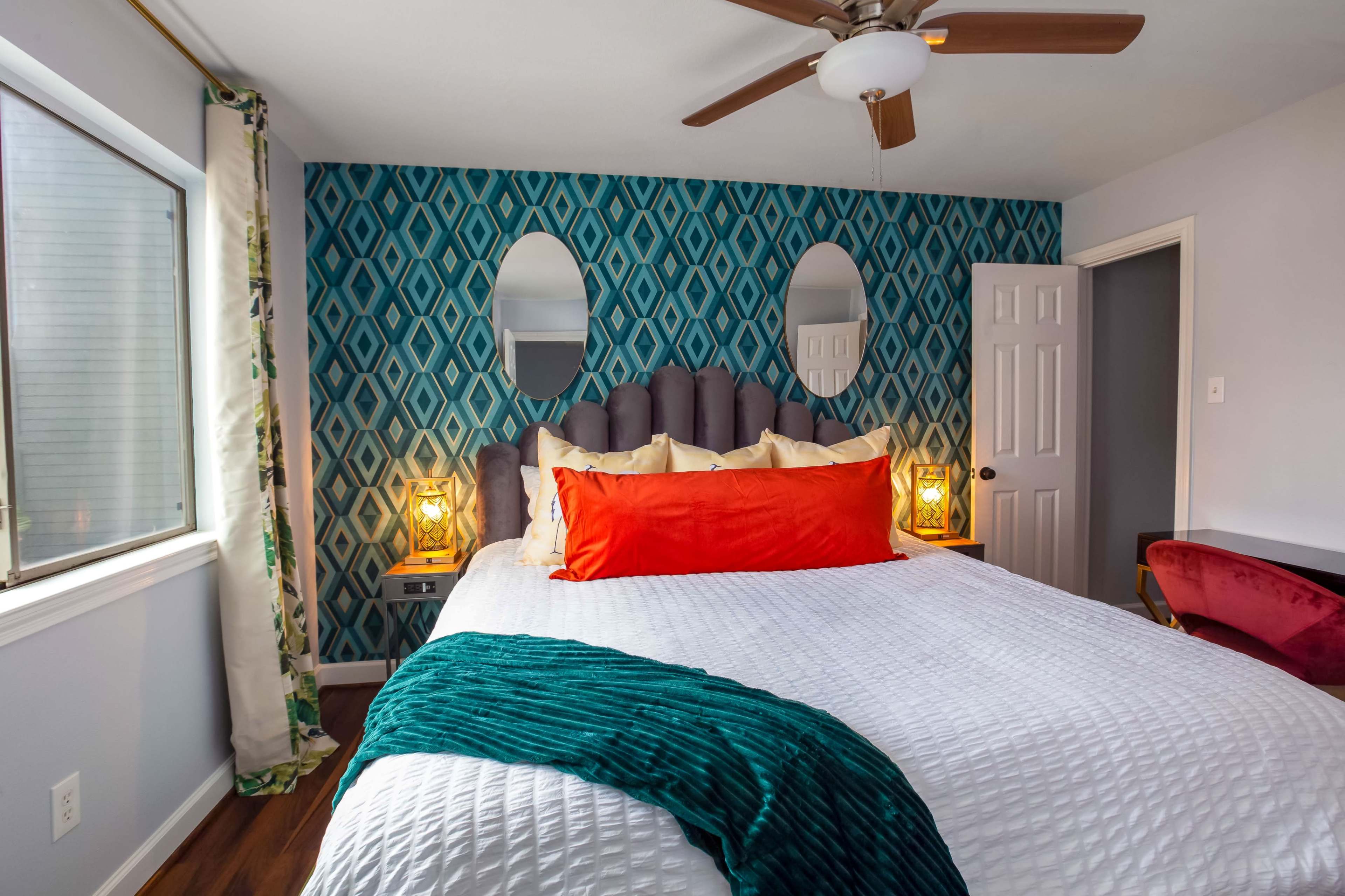 A bedroom featuring a large bed with an orange accent pillow, patterned teal wallpaper, and decorative lighting on either side of the bed.
