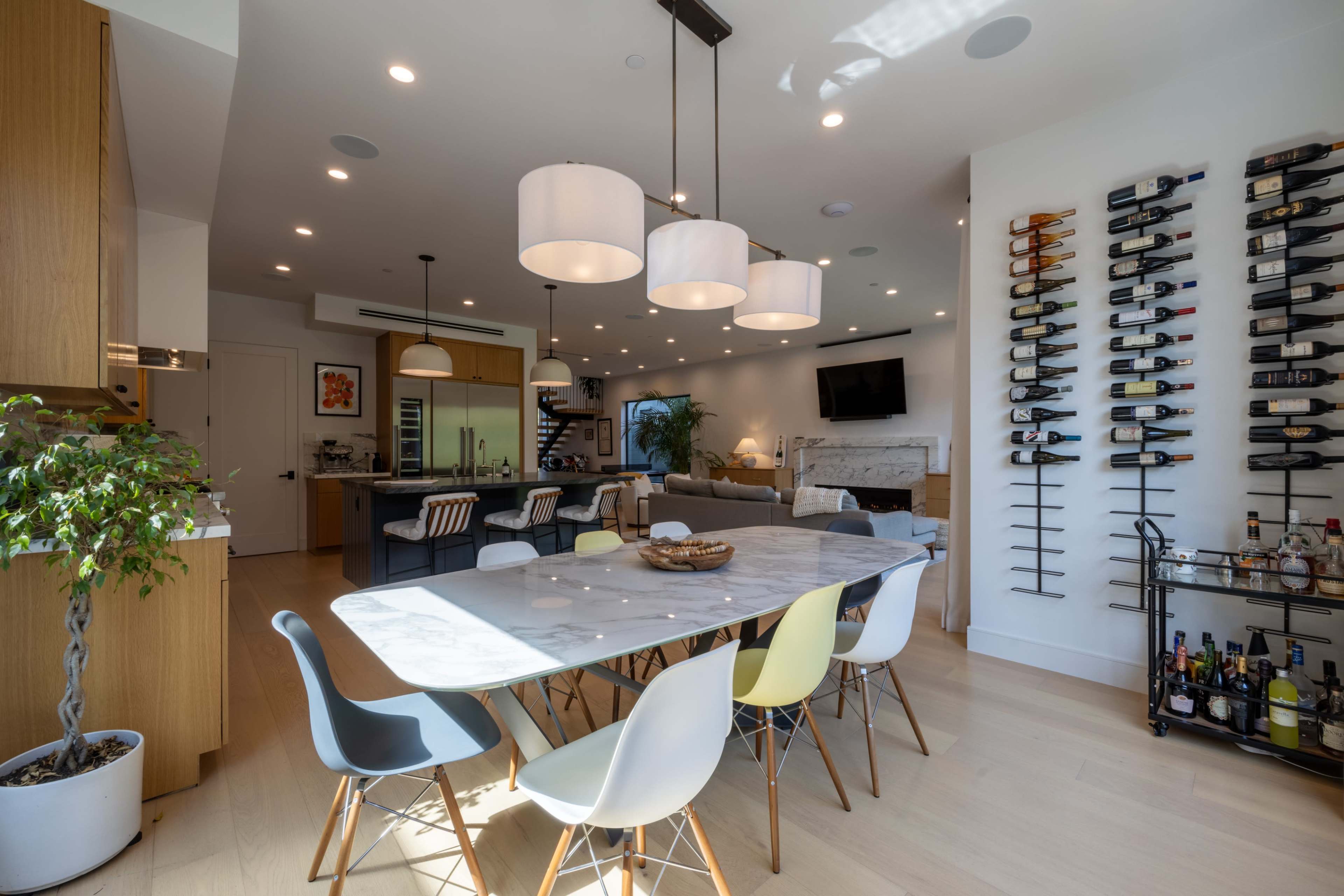 A modern open-concept kitchen and dining area featuring a marble dining table surrounded by colorful chairs, a wine rack on the wall, and a cozy seating area with a television.