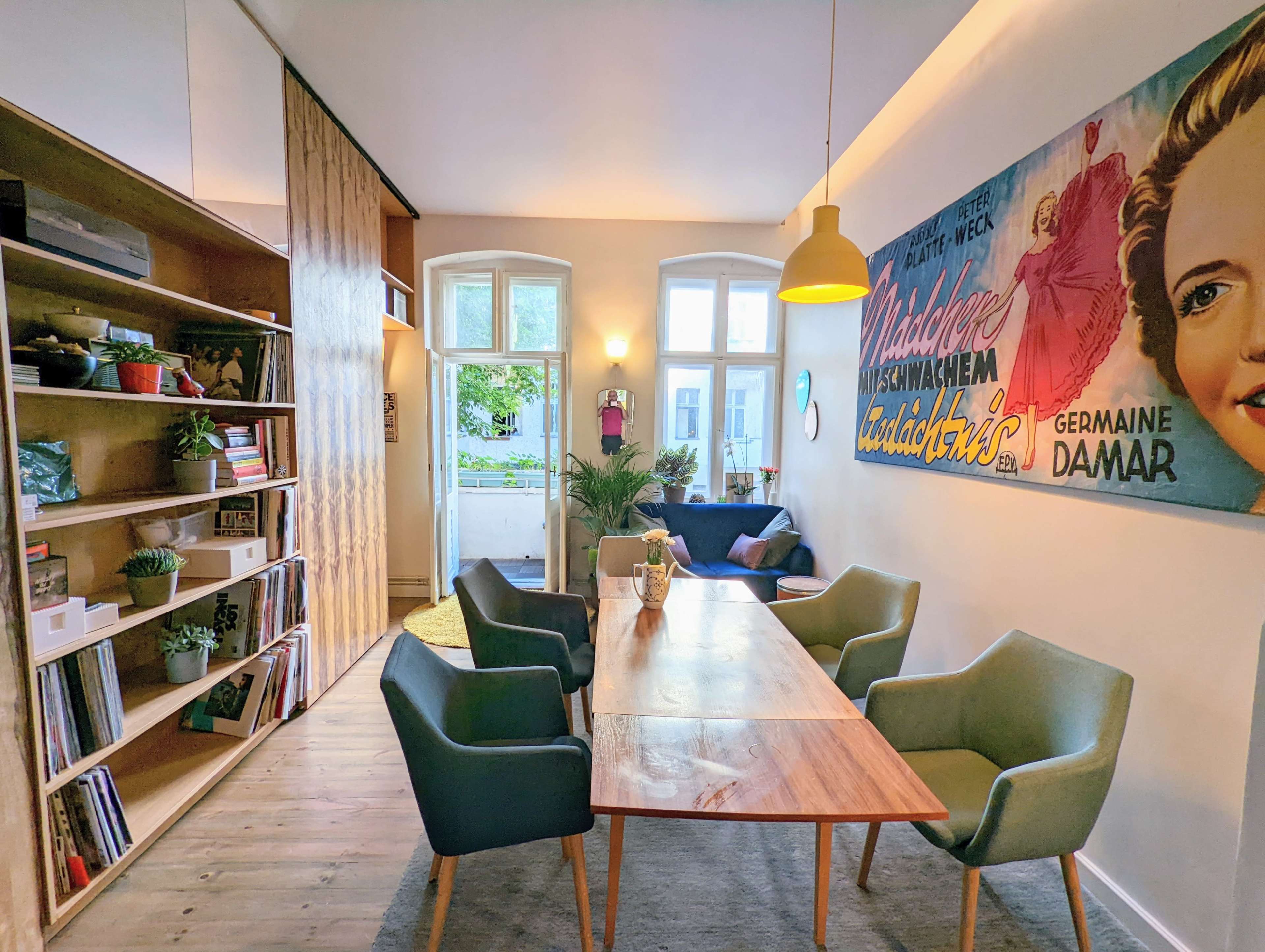 A modern living space with a wooden dining table surrounded by four chairs, a bookshelf filled with plants and books, and a vibrant wall adorned with a vintage poster.