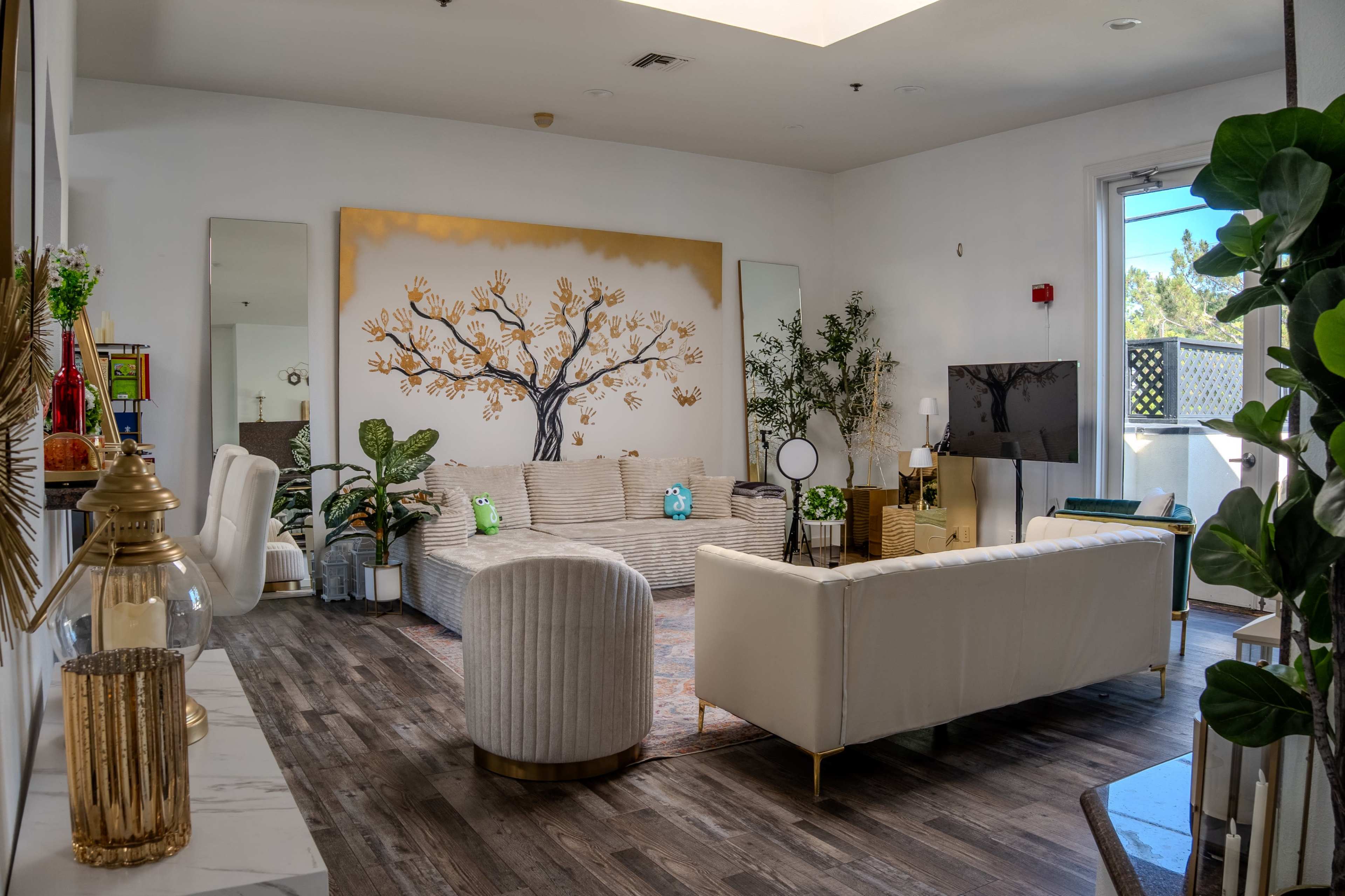 The image shows a bright, modern living room featuring a large wall mural of a tree, comfortable seating arrangements, and decorative plants.