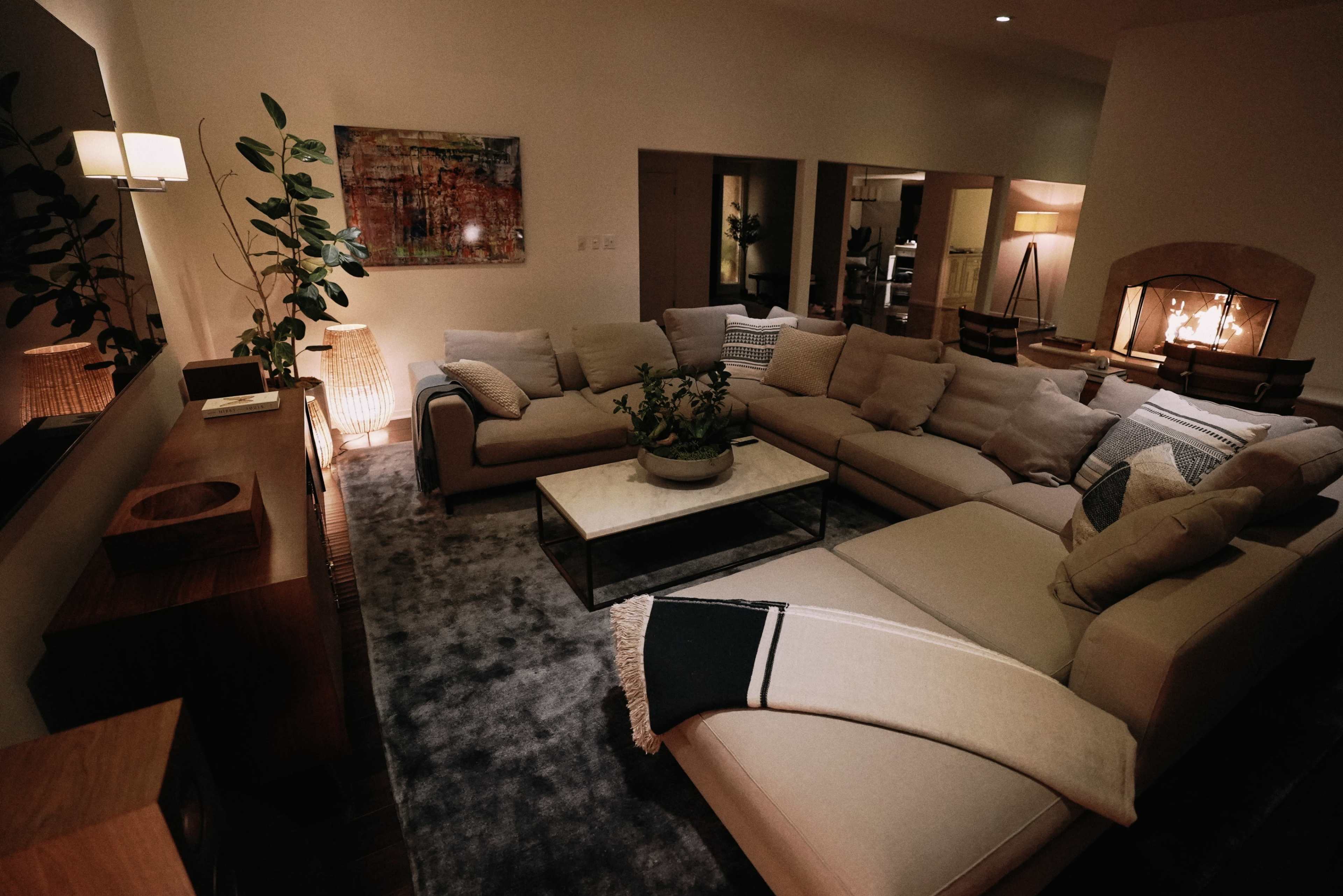 The image shows a modern living room featuring a large sectional sofa, a coffee table, indoor plants, and a fireplace.