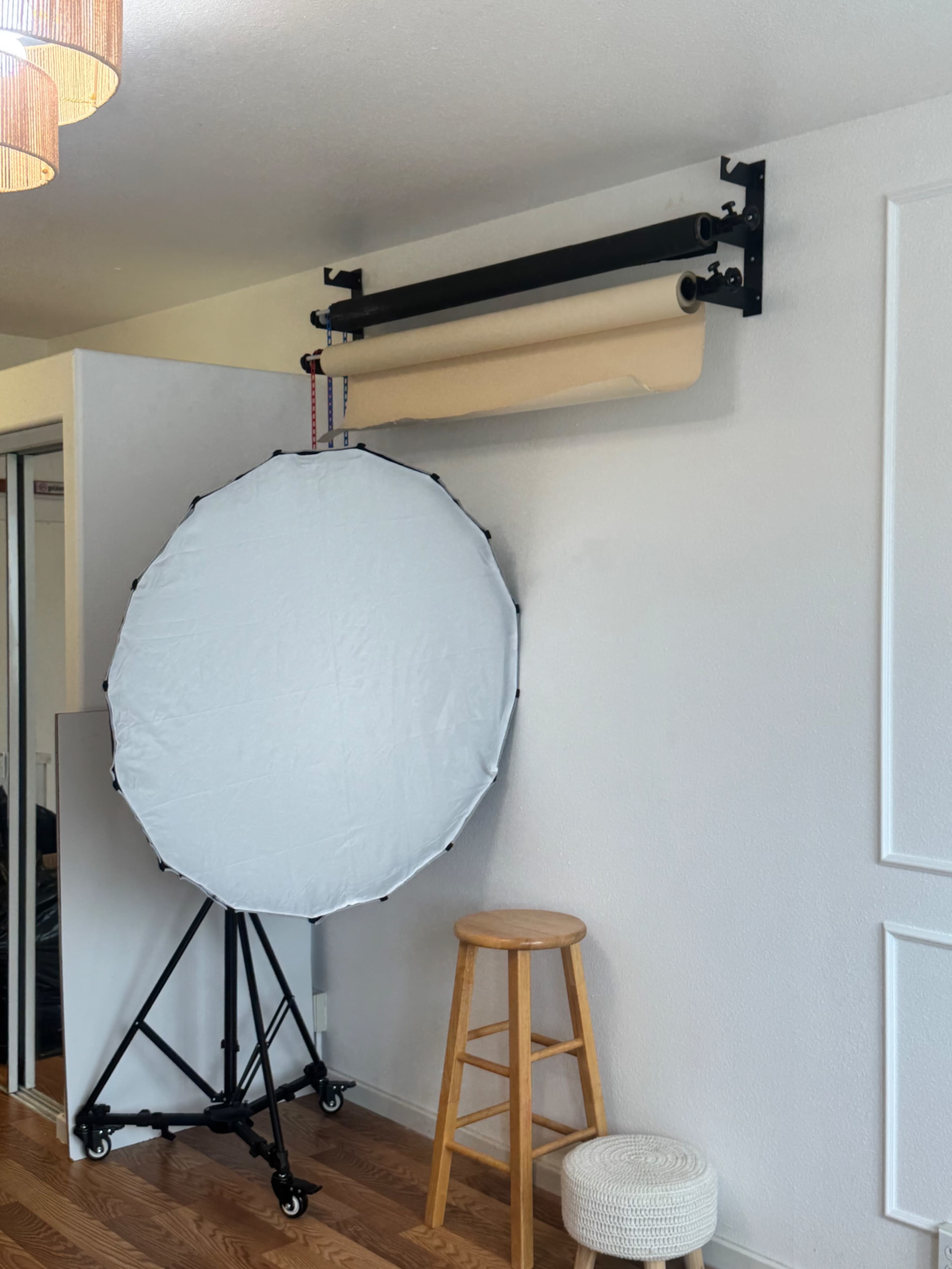 The image shows a photography setup featuring a large collapsible reflector, a stool, and a roll of backdrop paper mounted on a wall bracket.