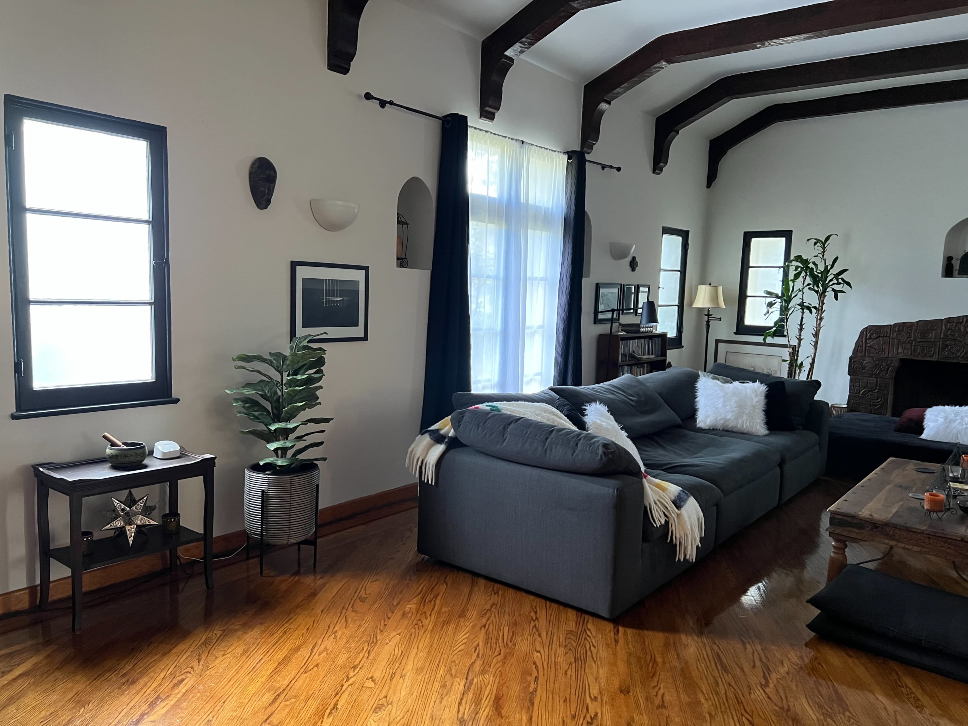 A spacious living room features a gray sofa, wooden beams on the ceiling, and a small table with a plant and decorative items.