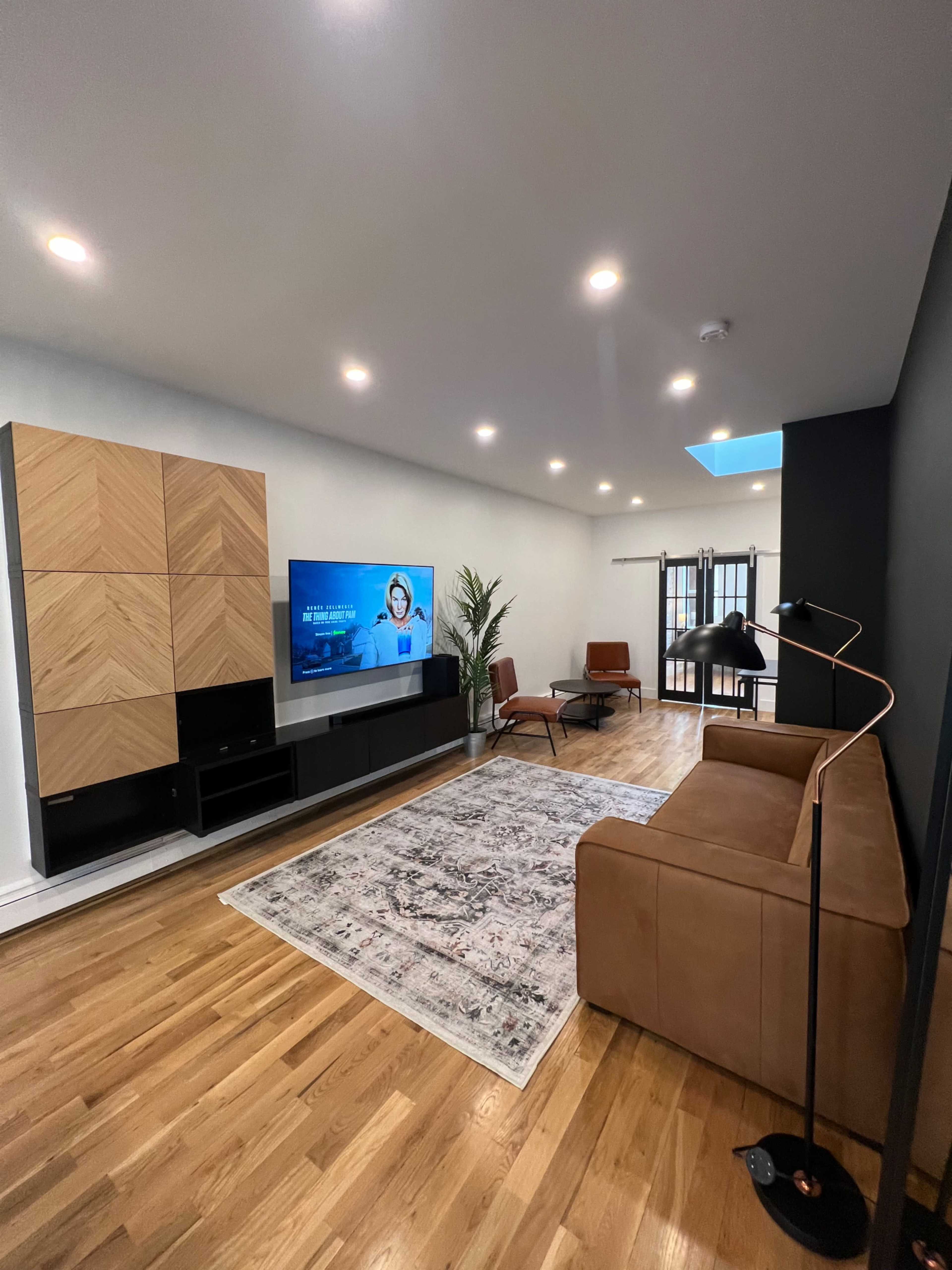 The living room features a brown leather sofa, a television mounted on a wooden cabinet, and a coffee table on a patterned rug, with large windows allowing natural light to enter.