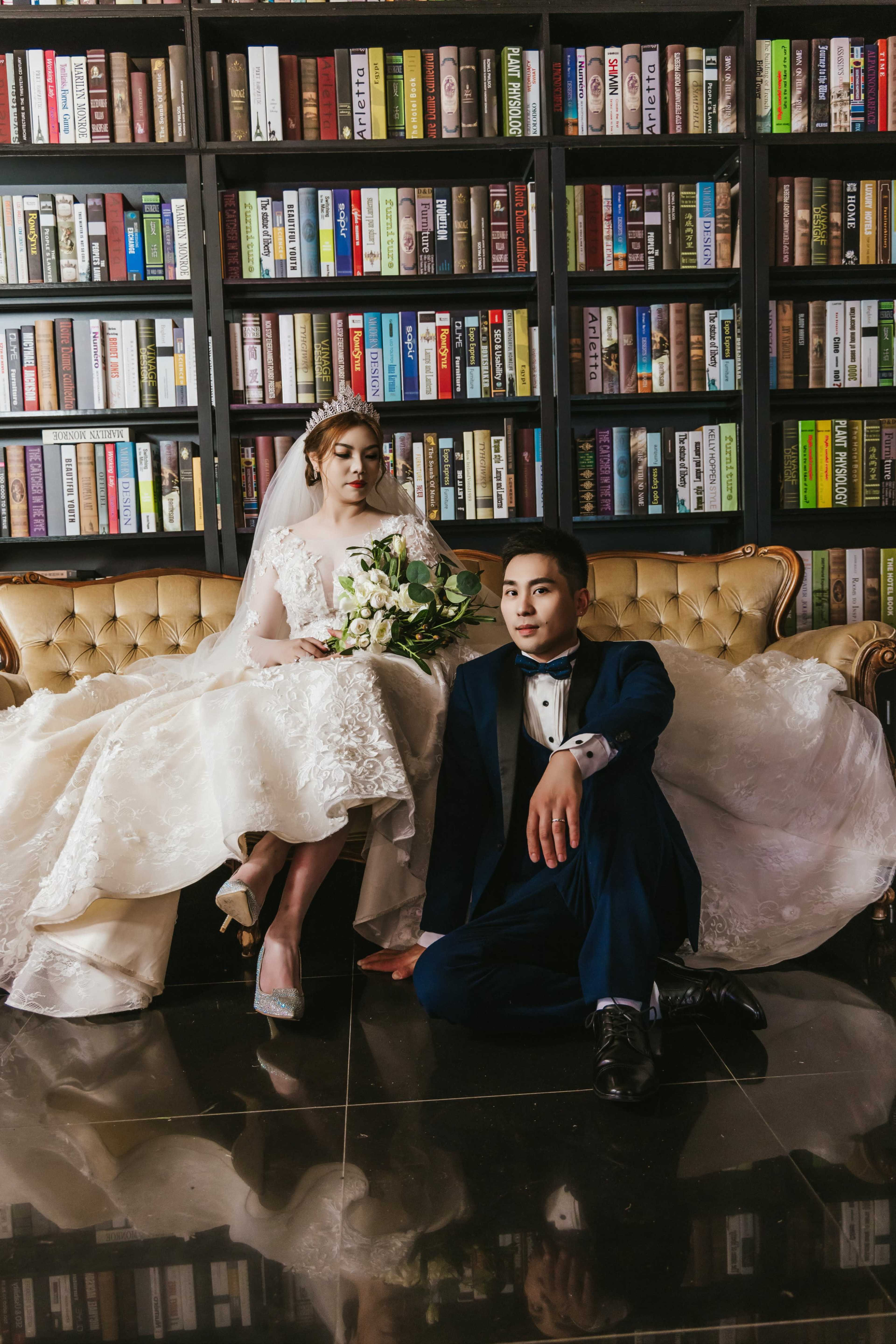 A bride in a white gown sits on a vintage sofa next to a groom in a blue tuxedo, surrounded by shelves filled with books.