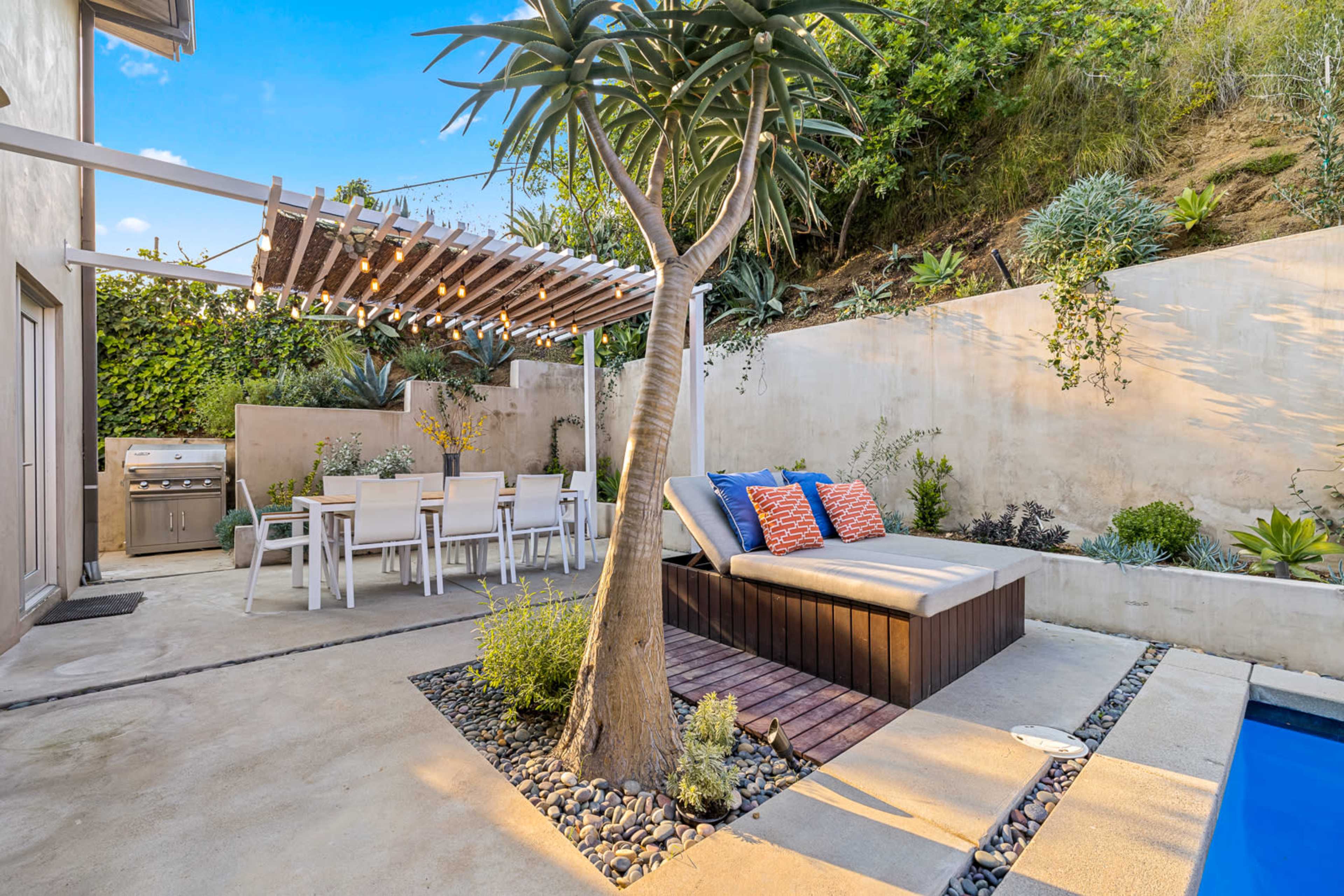 The image shows a modern outdoor patio area featuring a lounge space with pillows, a dining table, a grill, and a pool surrounded by greenery.