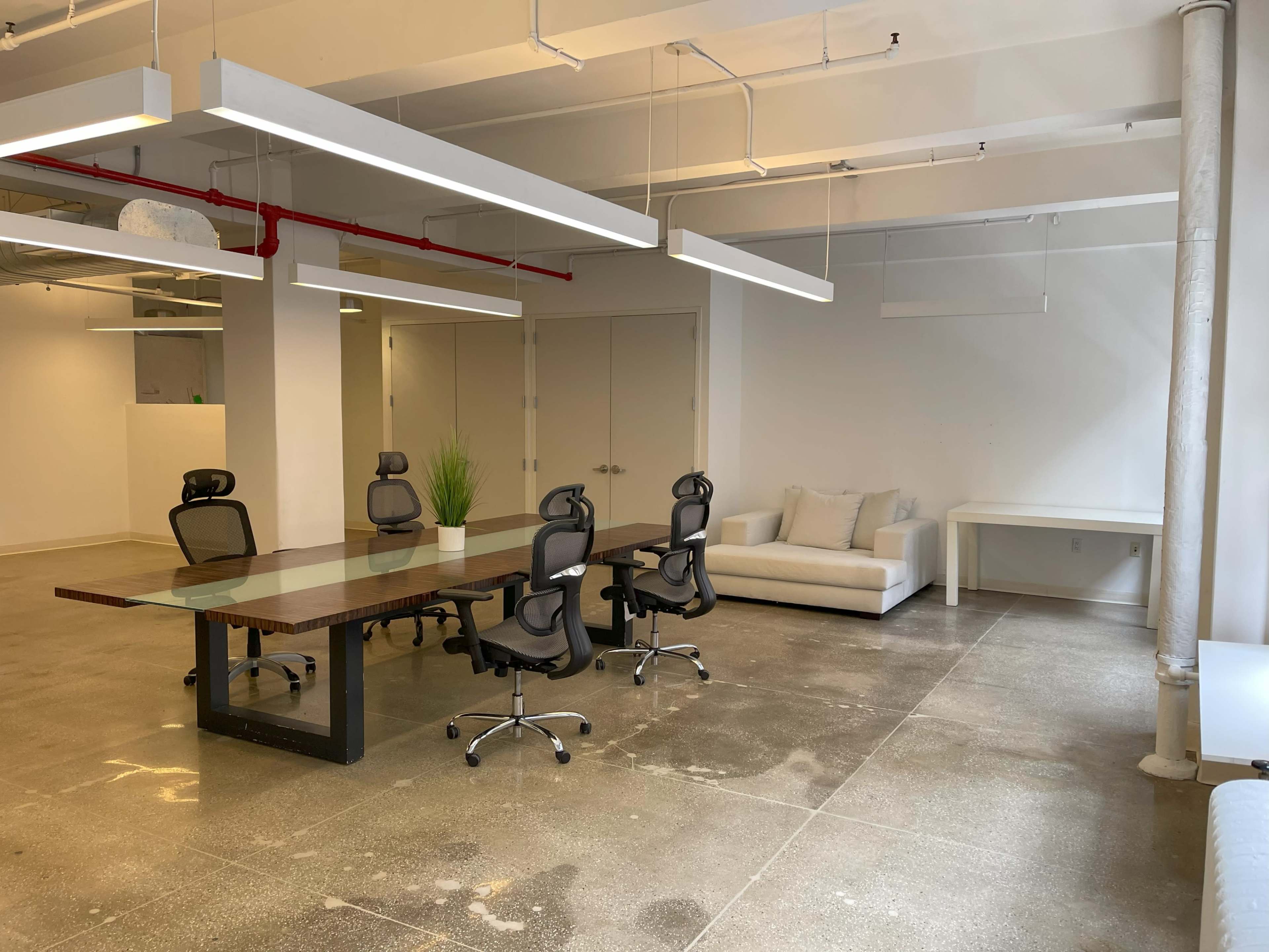A modern office space features a large glass table surrounded by four black ergonomic chairs, with a white sofa and potted plant against a white wall.