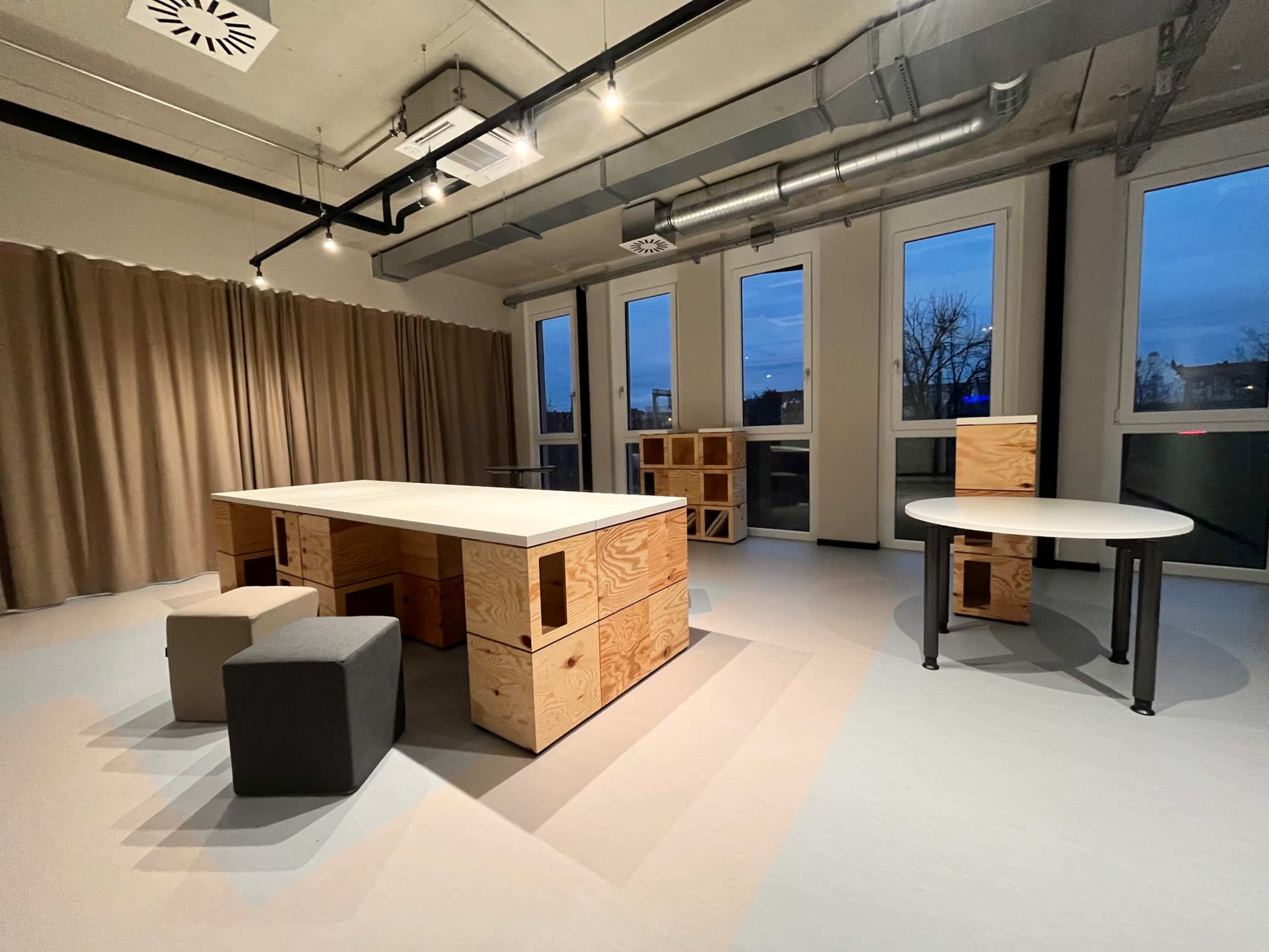 The image shows a spacious room with large windows, featuring a table made of wooden boxes, a round table, and two stools in a minimalist design.