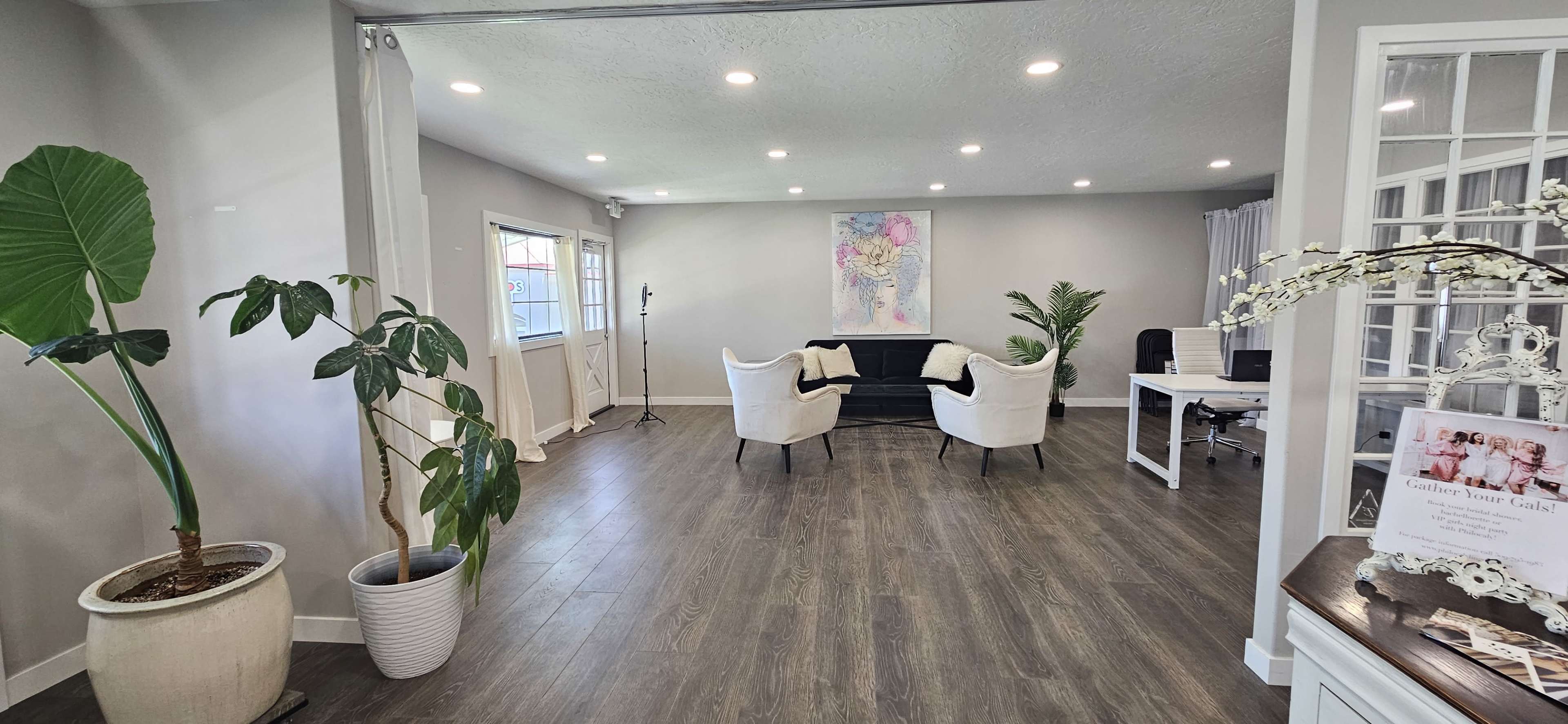 A modern, open room with a seating area featuring two white chairs and a black couch, along with plants and a desk in a well-lit environment.