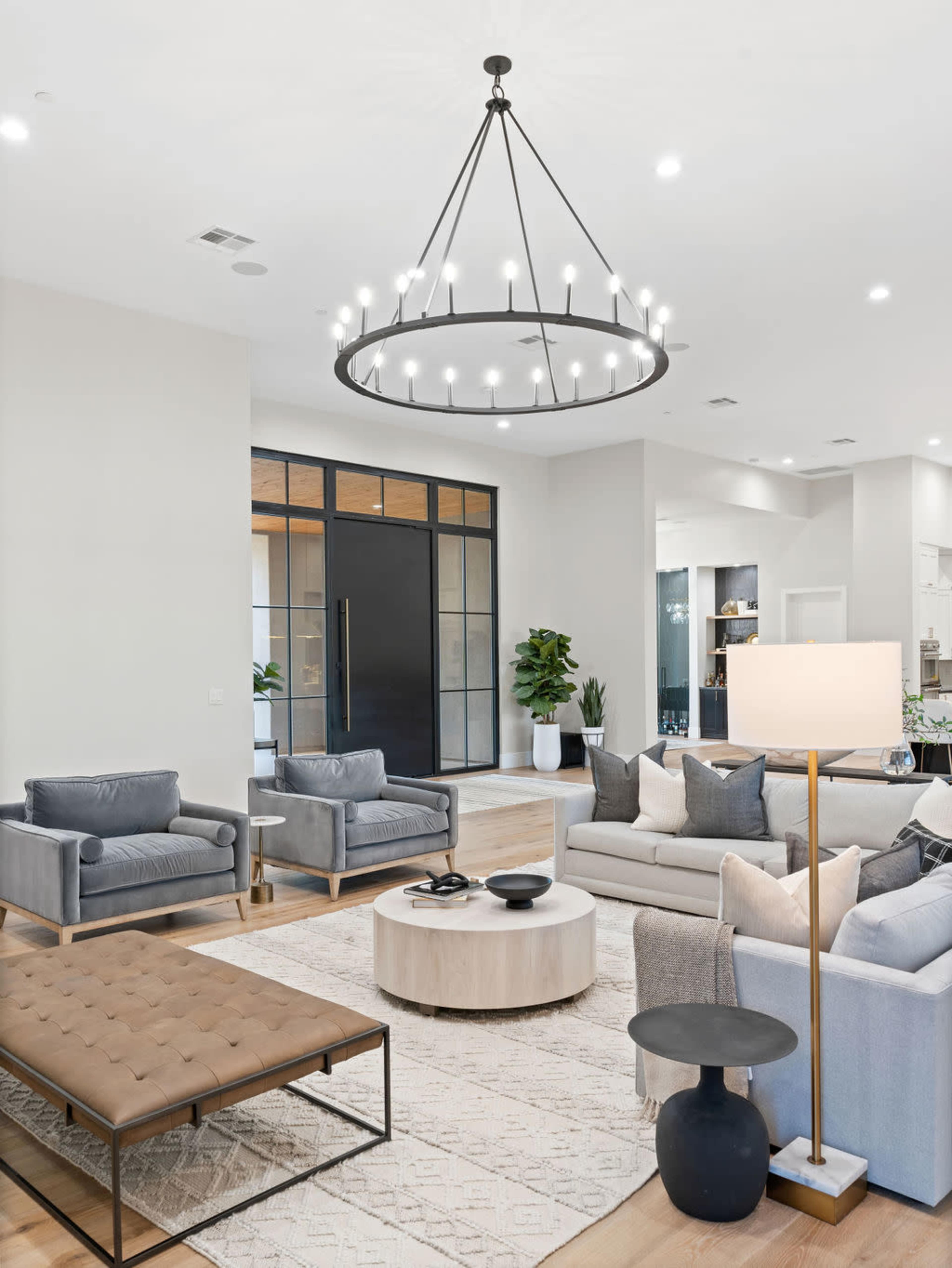 A modern living room features gray upholstered furniture, a round coffee table, and a large chandelier above, with glass doors leading to another area.