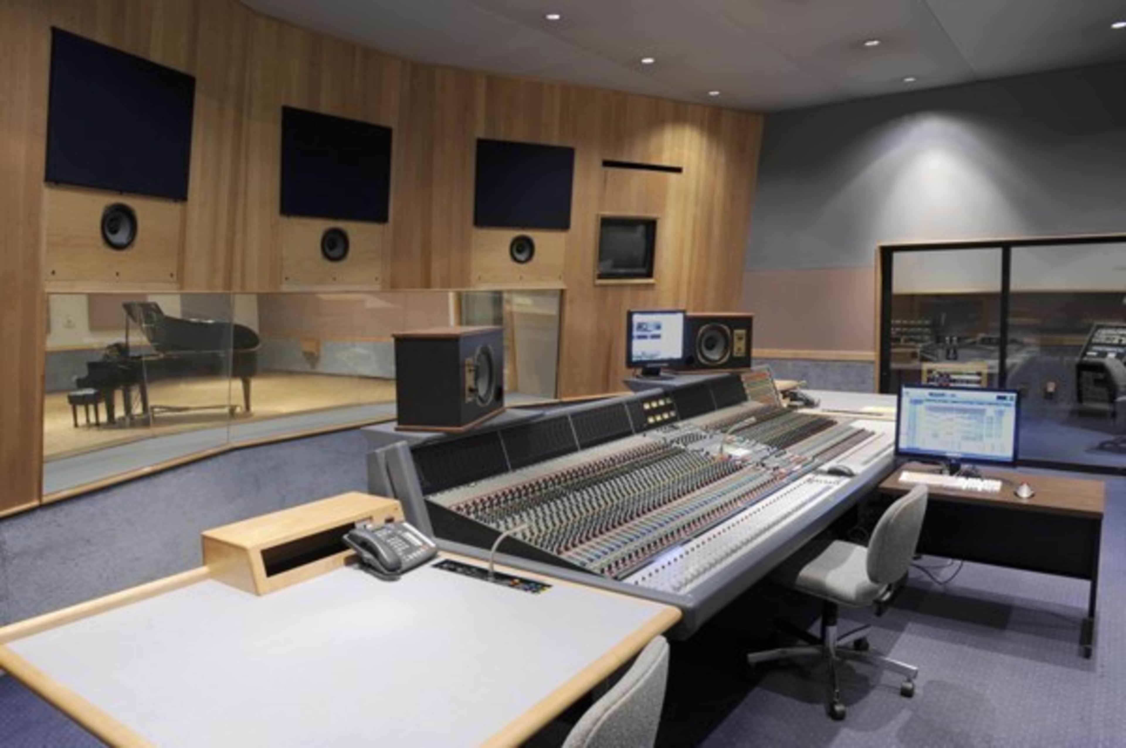 A modern recording studio featuring a large sound mixing console, several computer monitors, and a glass-walled area containing a grand piano.