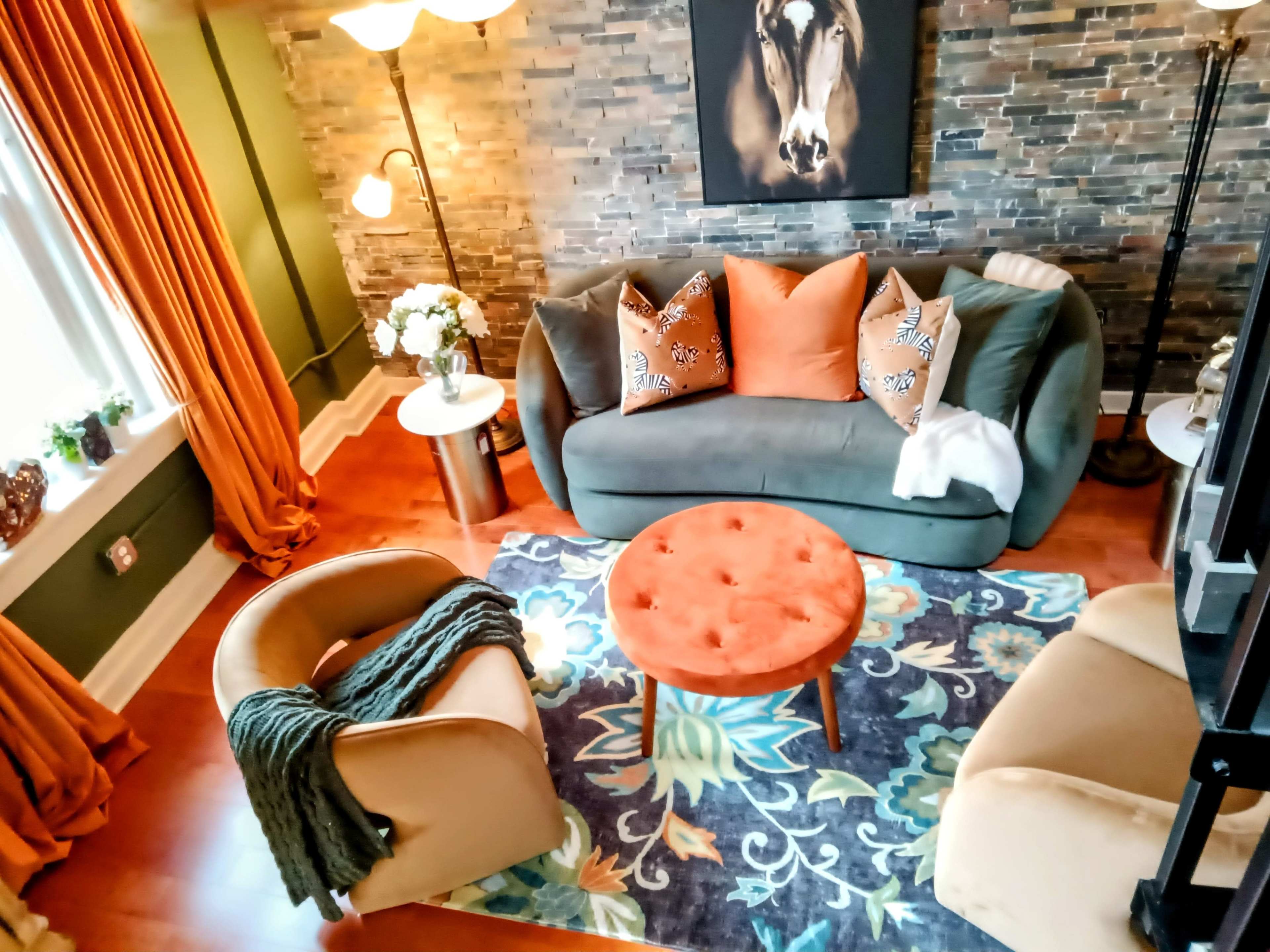 A cozy living room featuring a green sofa, orange accent chairs, a round orange coffee table, and a colorful rug with floral patterns.