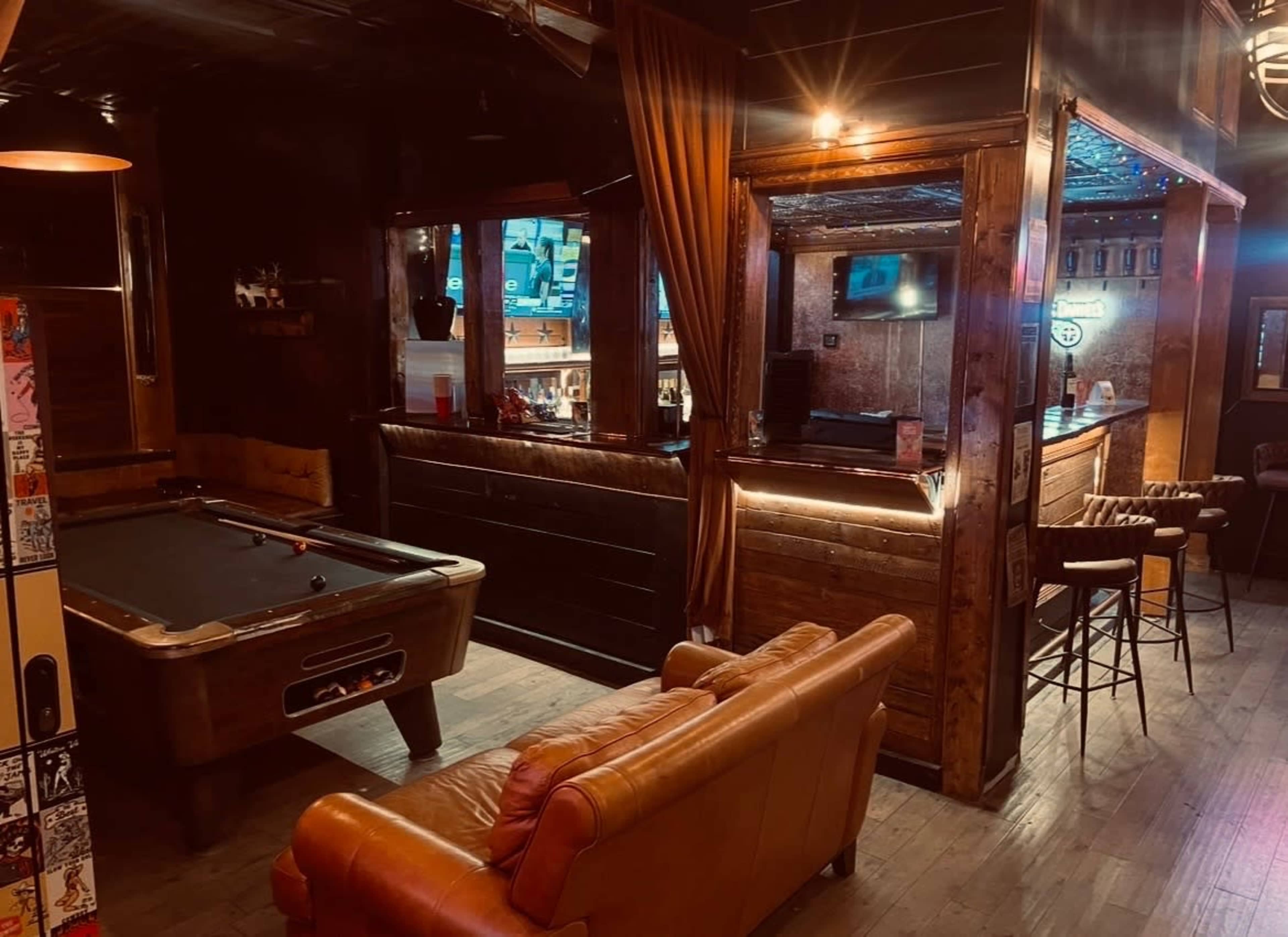 The image shows a dimly lit bar area featuring a pool table, a leather sofa, and a wooden bar counter with stools.