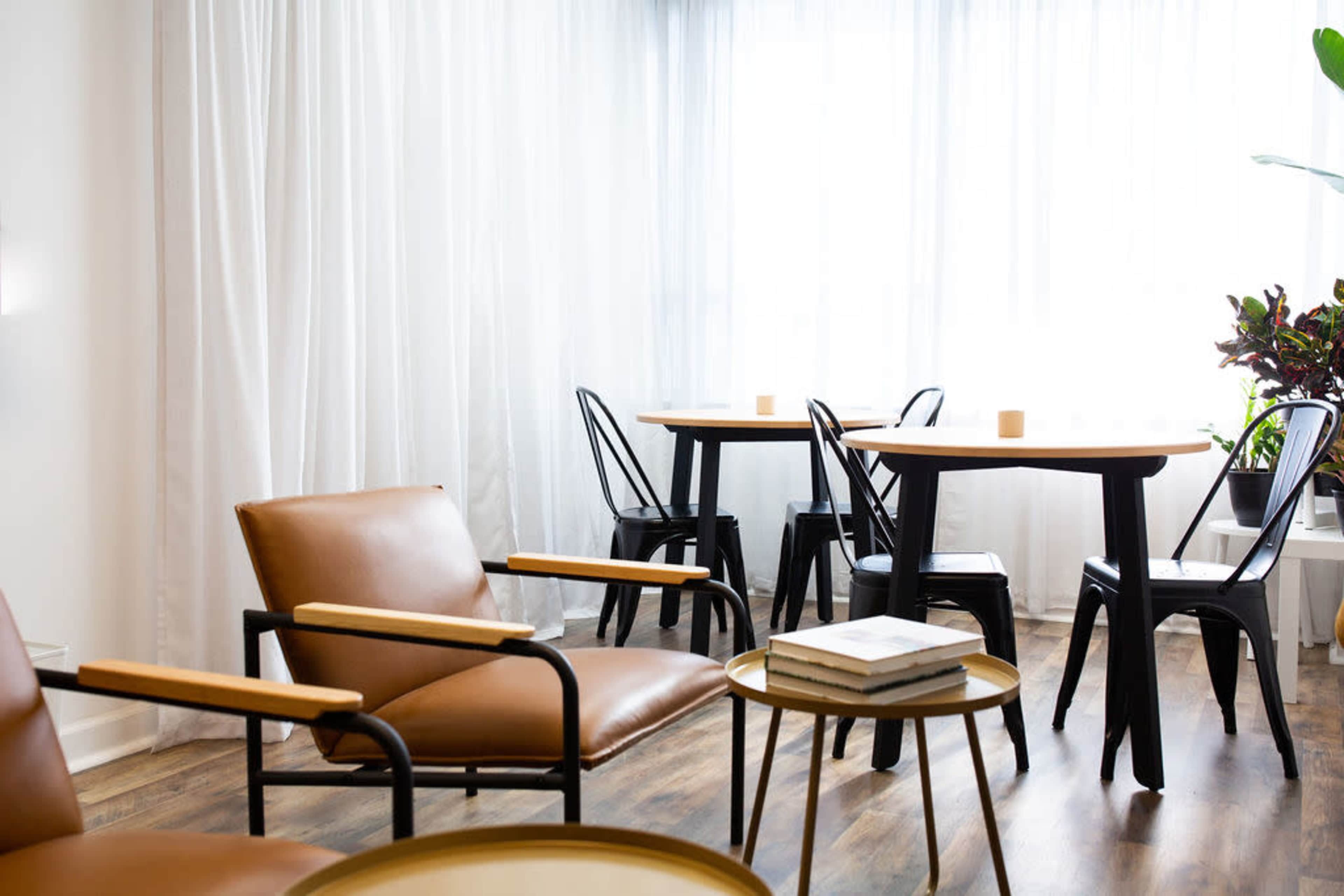 A bright, minimalist room featuring brown leather chairs, black metal tables, and large windows dressed with sheer white curtains.