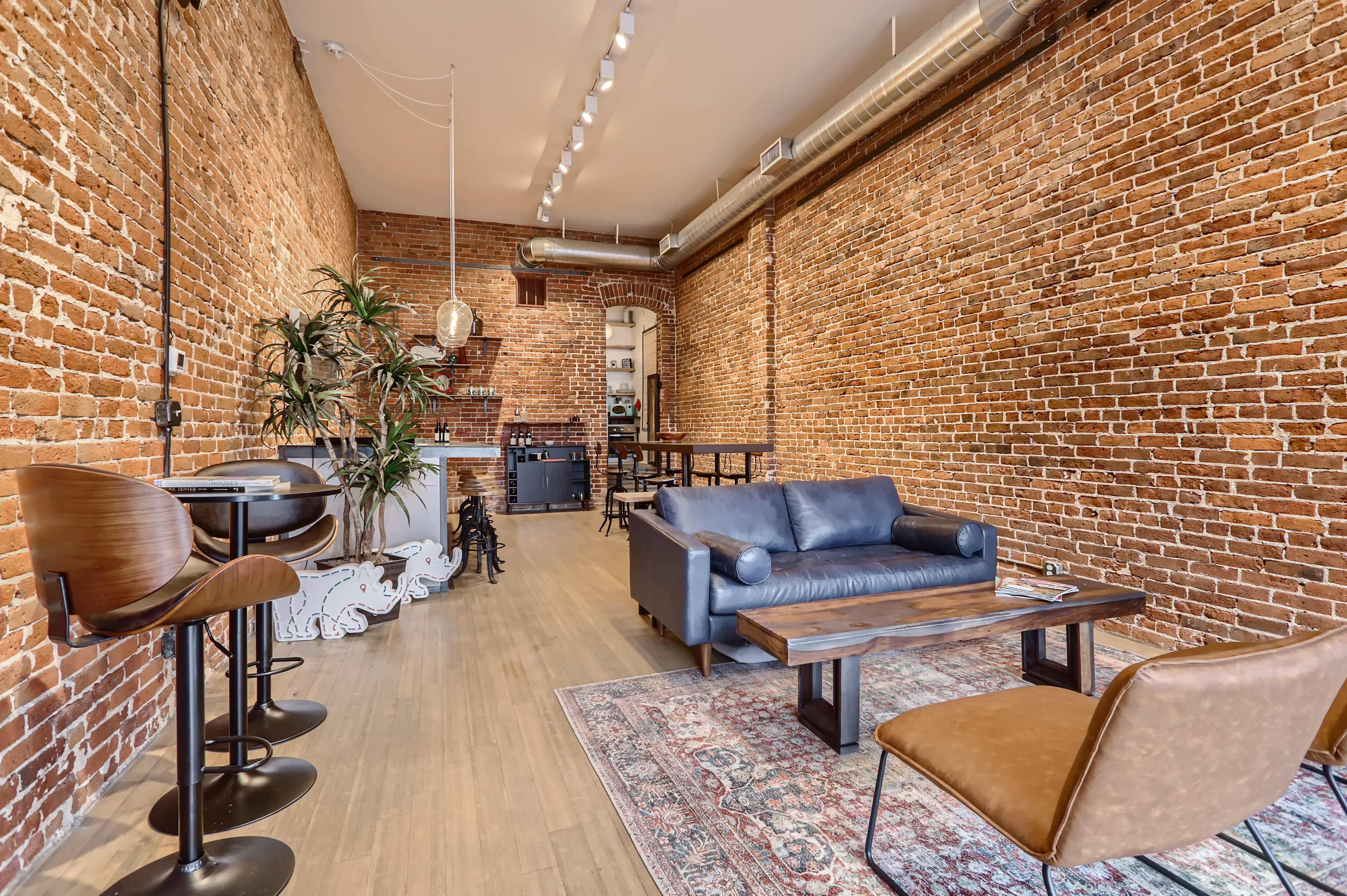 A spacious interior with exposed brick walls, hardwood flooring, and a mix of seating areas including a leather sofa, a coffee table, and high-backed bar stools at a counter.