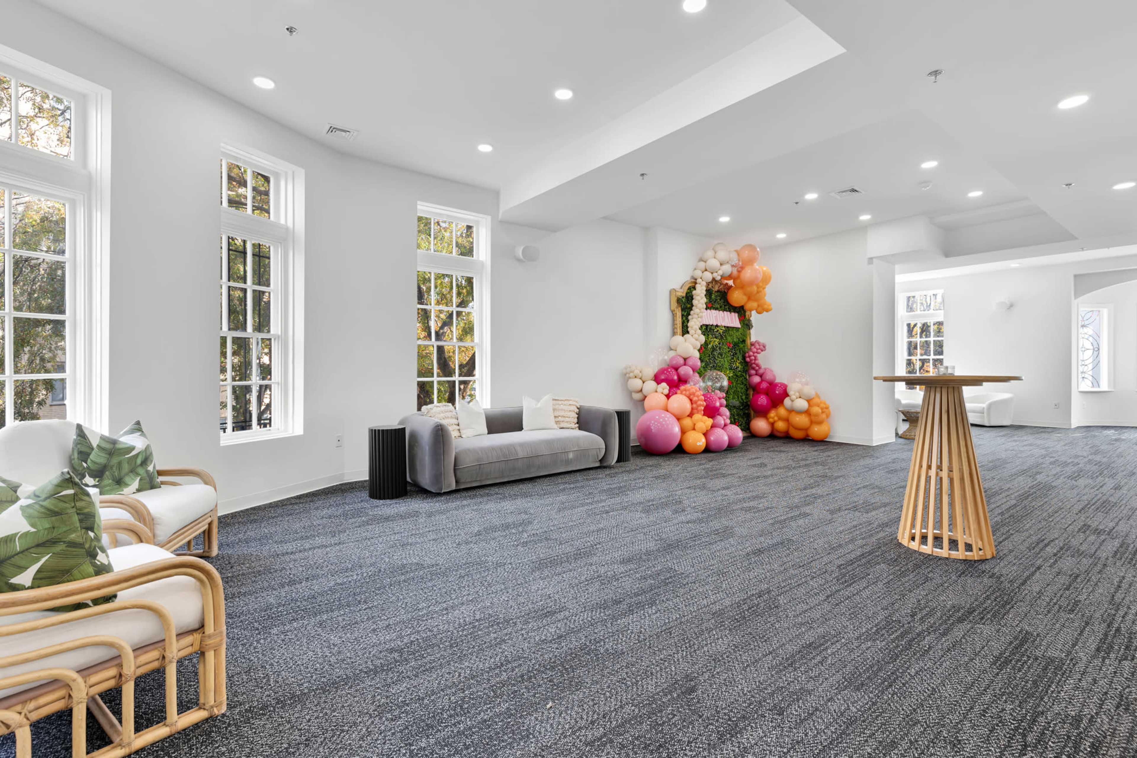 A spacious, well-lit room features large windows, a gray sofa, a wooden table, and a decorative arrangement of colorful balloons against one wall.