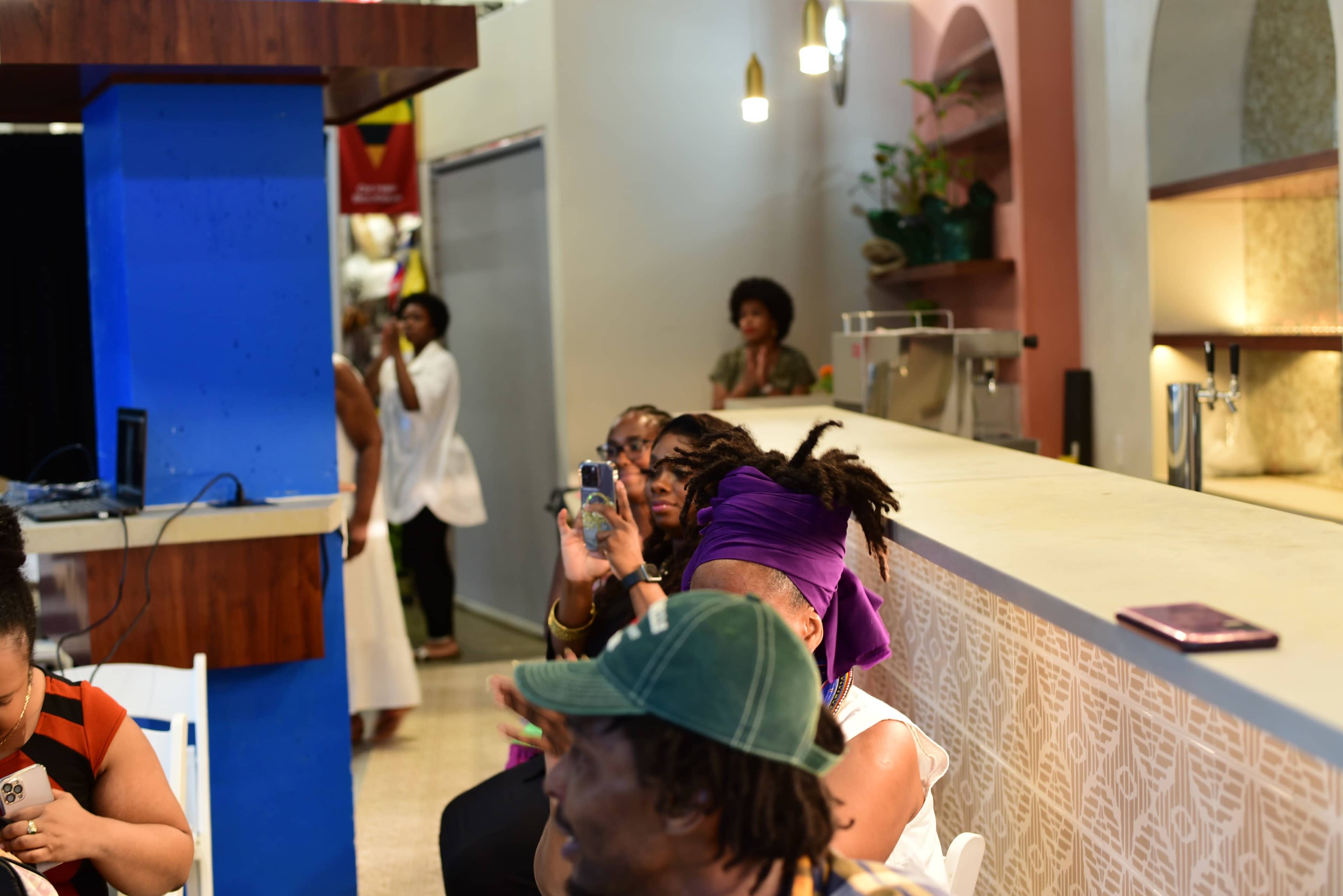 A group of people are seated indoors, with some taking photos and a few others engaged in conversation, while a brightly decorated bar area is visible in the background.
