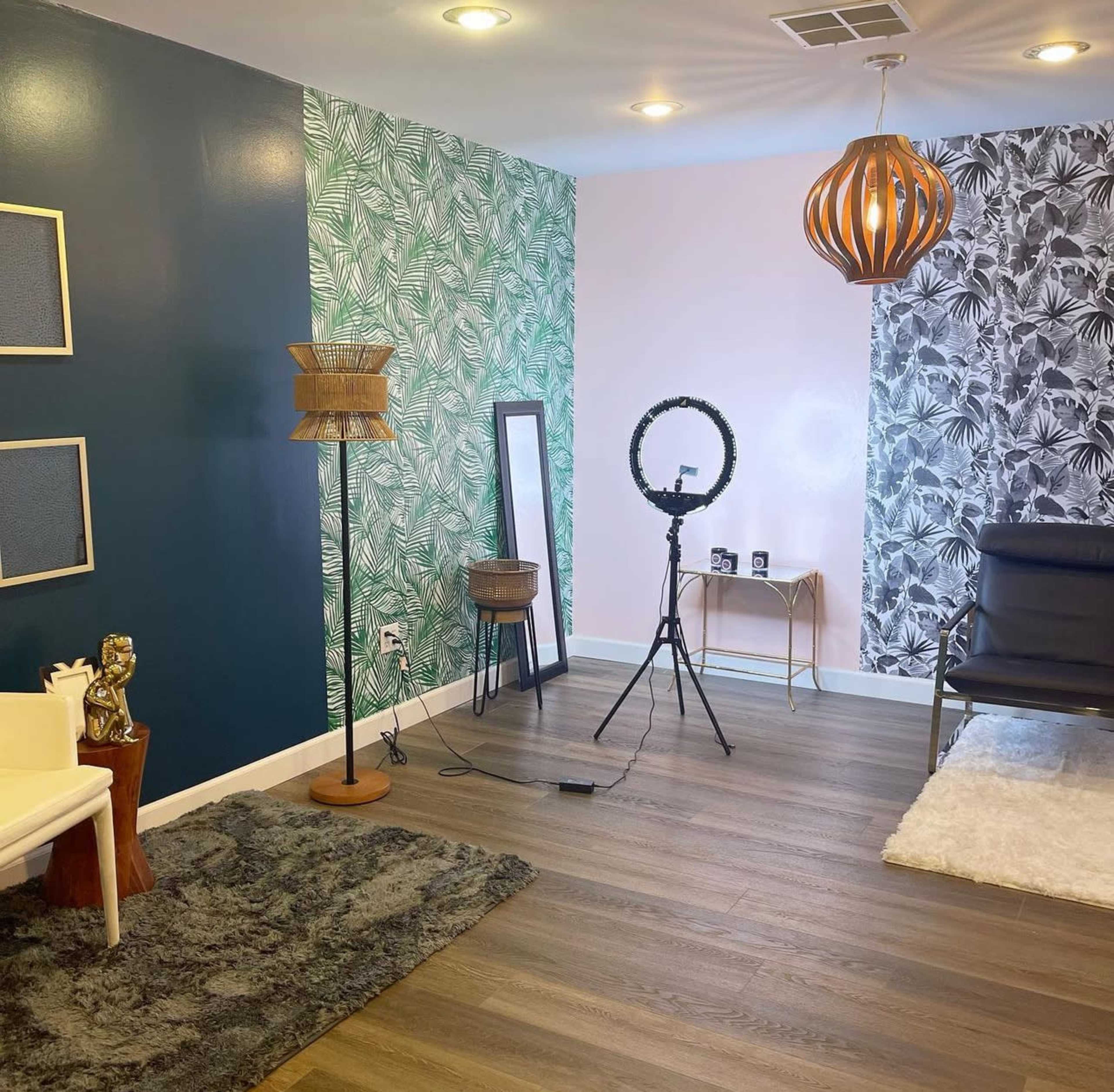 The image shows a well-decorated room featuring patterned wallpaper, a floor lamp, a mirror, a ring light on a tripod, and a modern chair on wooden flooring.